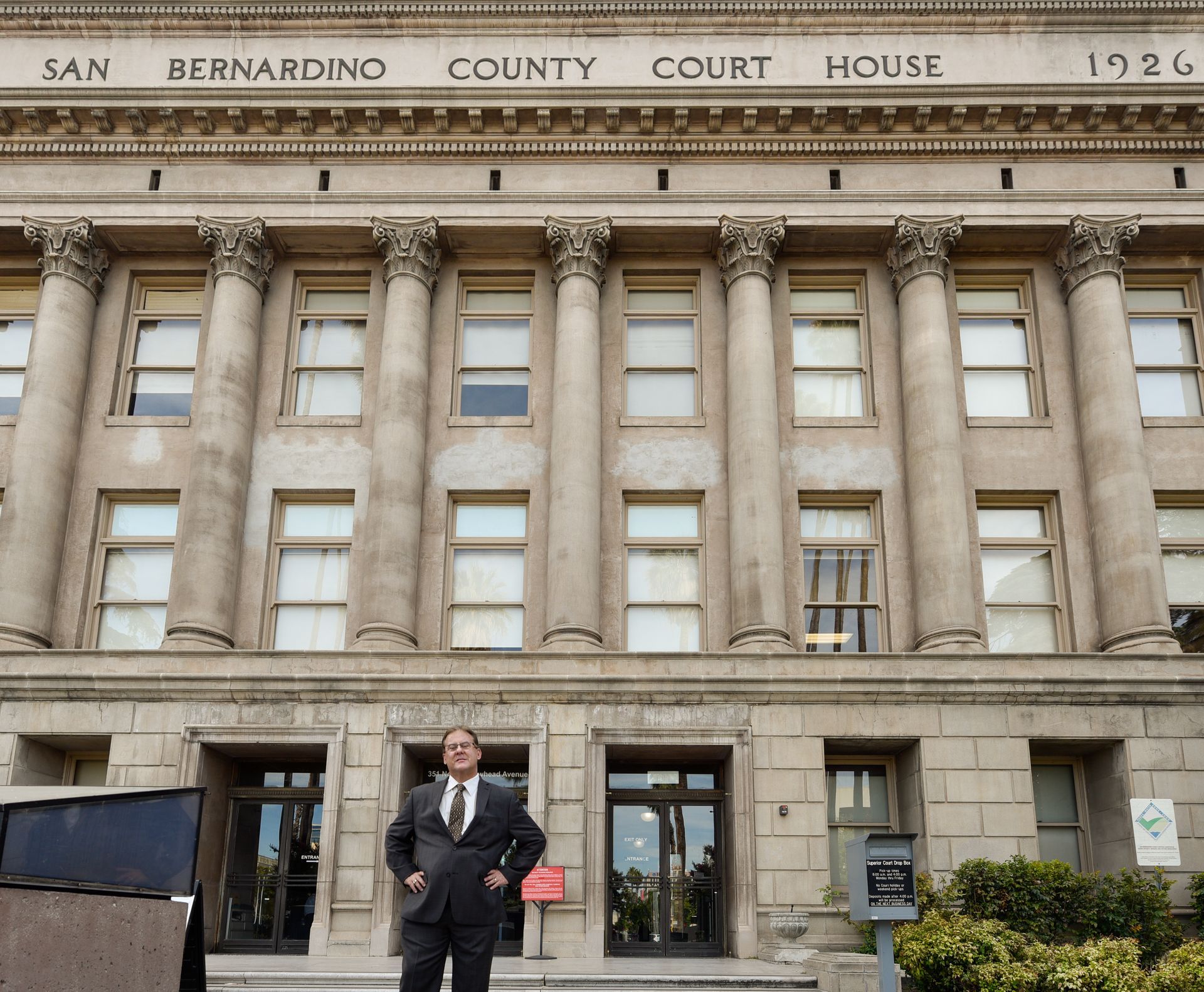A person in a suit stands with hands on hips in front of the historic 1926 San Bernardino County Courthouse.