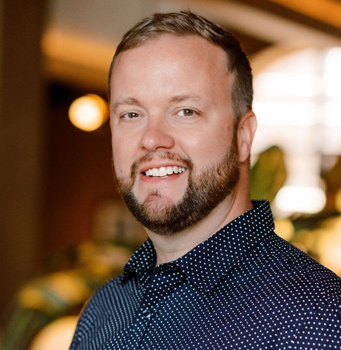 Smiling man with a beard in a navy polka-dot shirt, standing indoors with a blurred warm background