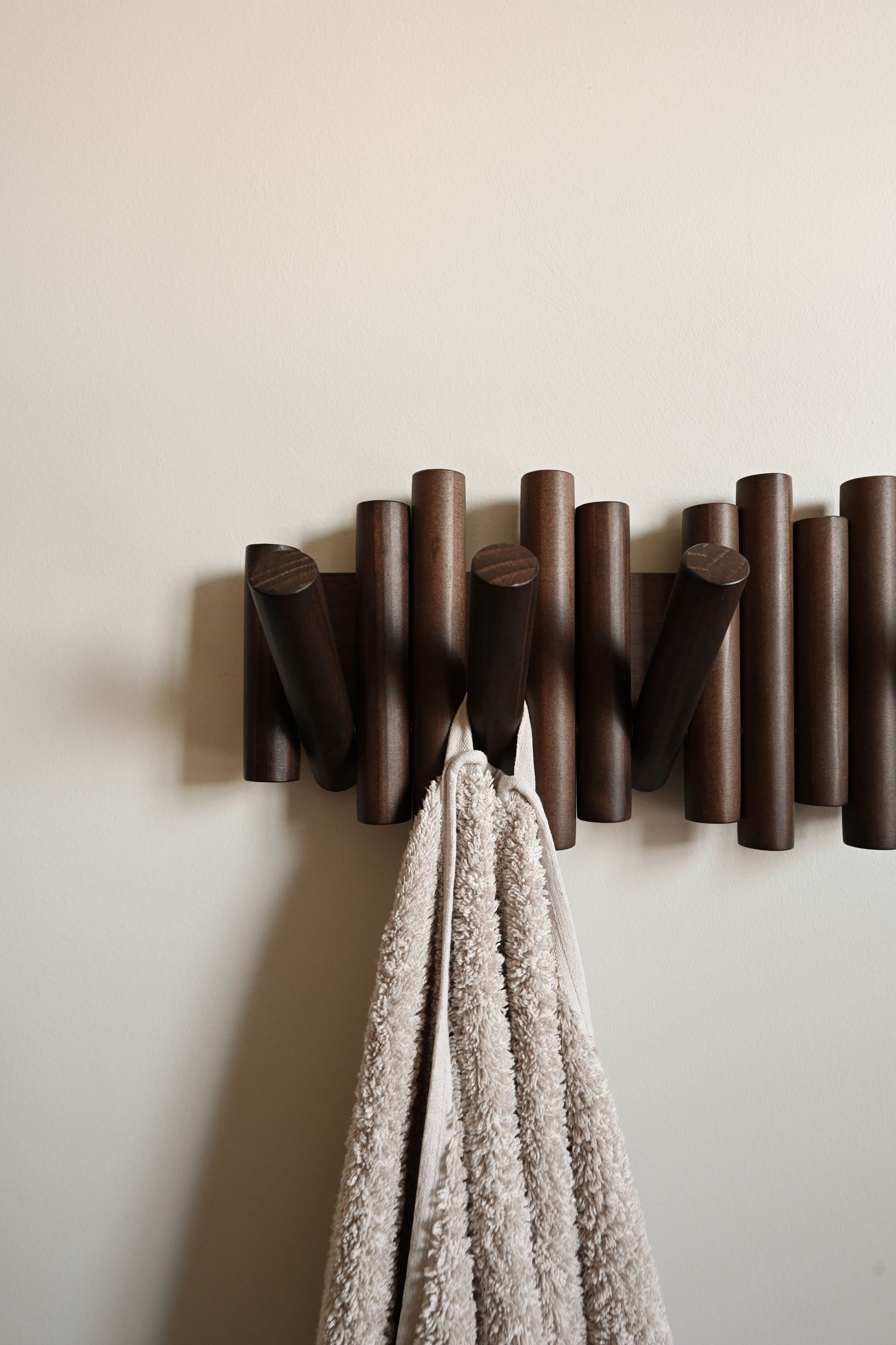 Houten wandkapstok met cilindrische haken, waaraan een opgevouwen beige handdoek tegen een lichte muur hangt.