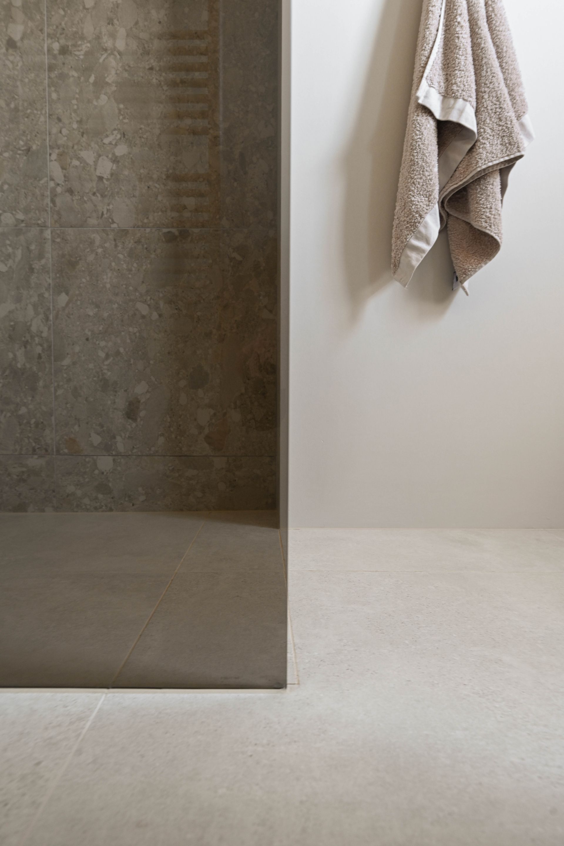 Grijze tegelwand in de douche grenst aan een gebroken witte muur met handdoek. Lichtgrijze vloer.
