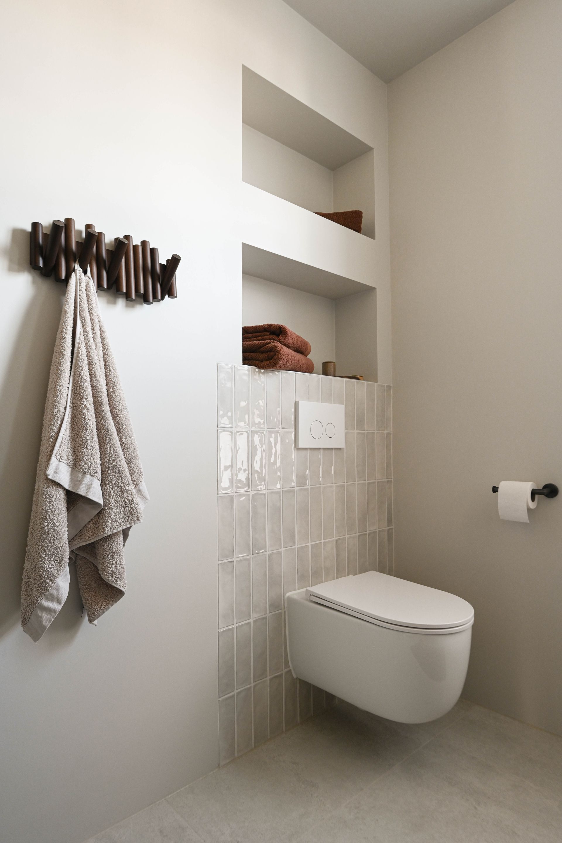 Moderne badkamer met wandtoilet, ingebouwde plankjes voor handdoeken en een handdoek die aan een houten wandhaak hangt.