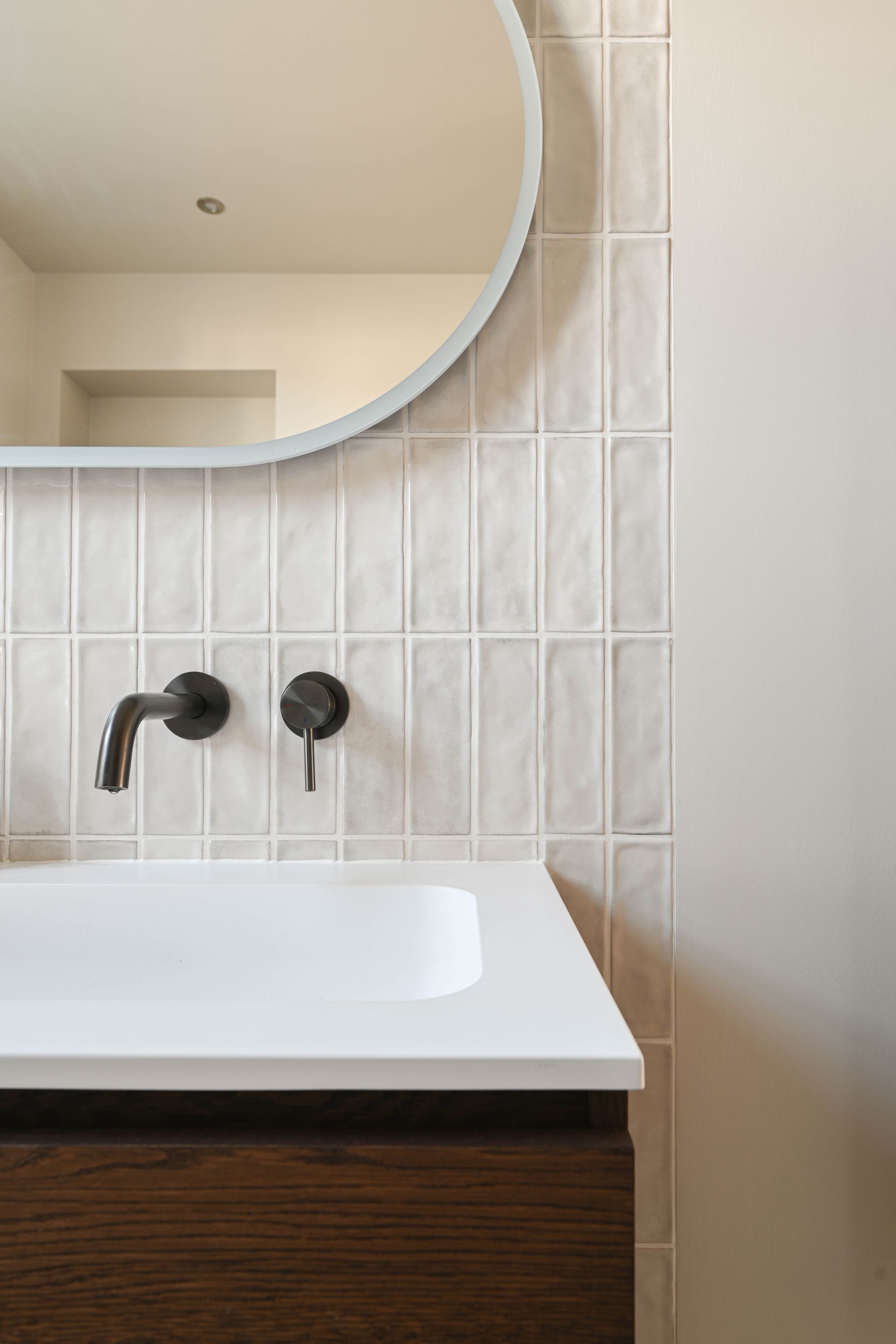 Badkamer met witte wastafel, donkere kranen, houten kast en een lichtgekleurde, verticaal betegelde achterwand onder een ronde spiegel.