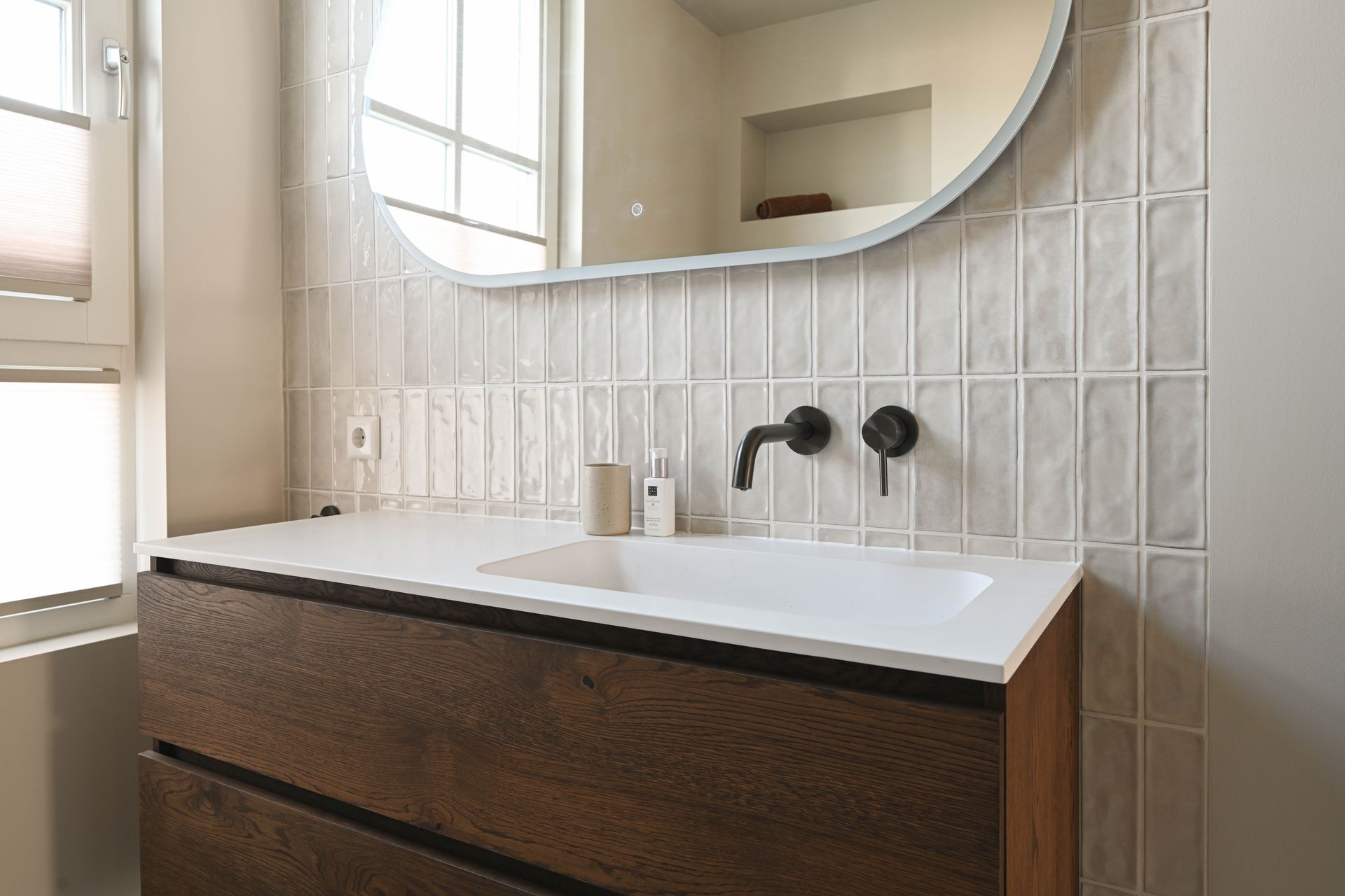 Badkamer met houten badmeubel, witte wastafel, matzwarte kraan en verticale tegels.