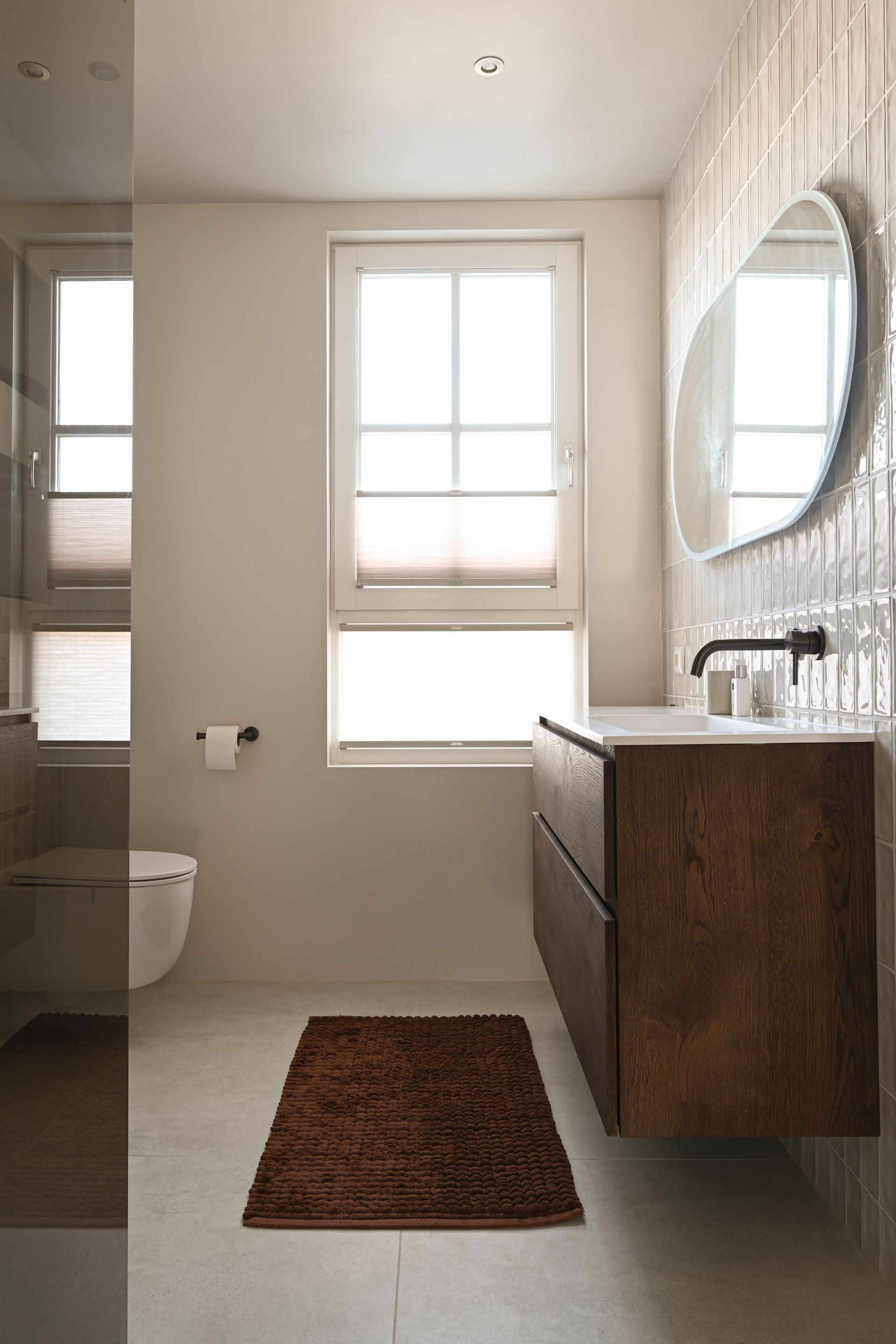 Moderne badkamer met bruin badmeubel, vloerkleed en een ovale spiegel. Wit toilet en witte jaloezieën, met een wand bekleed met verticale tegels.