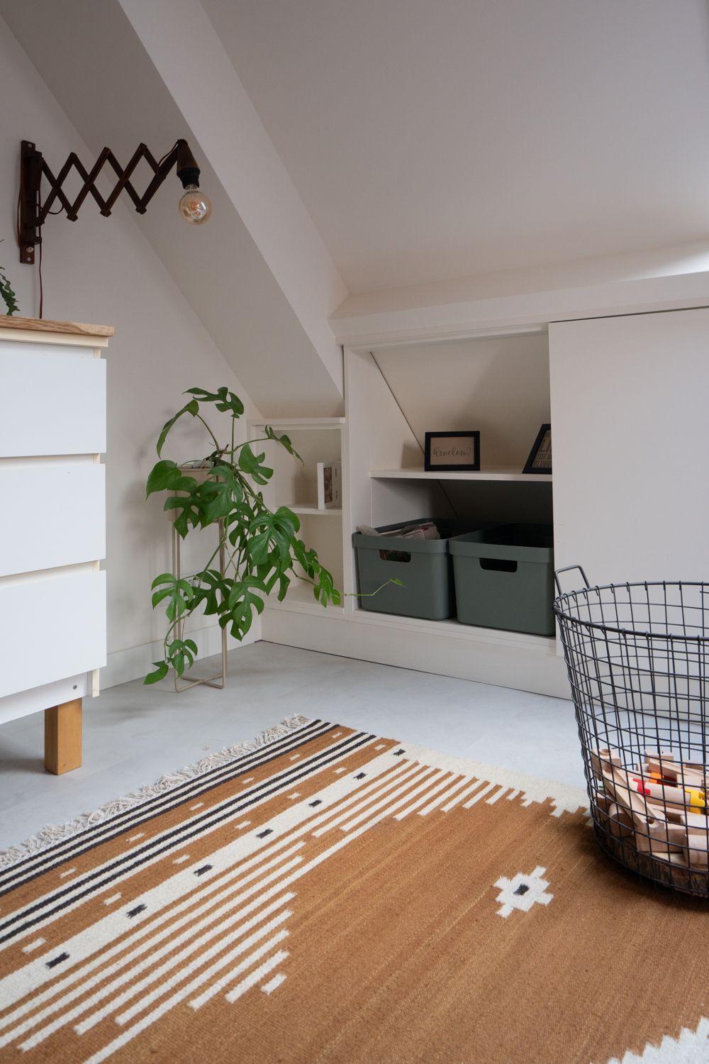 Gezellige zolderkamer met witte muren, ladekast, vloerkleed met patroon en een potplant.