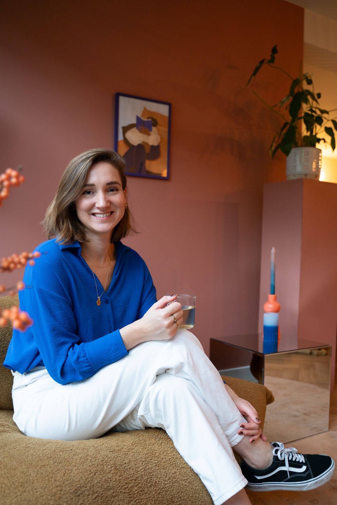 Een vrouw in een blauwe blouse en witte broek zit lachend op een bank.
