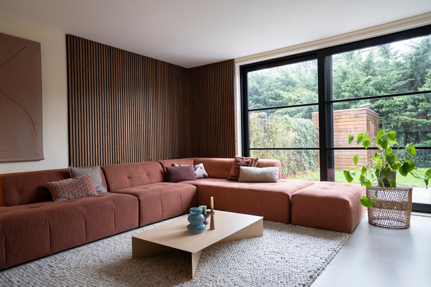 Gezellige woonkamer met roestkleurige hoekbank, houten wanddecoratie en een groot raam met uitzicht op een weelderige tuin.