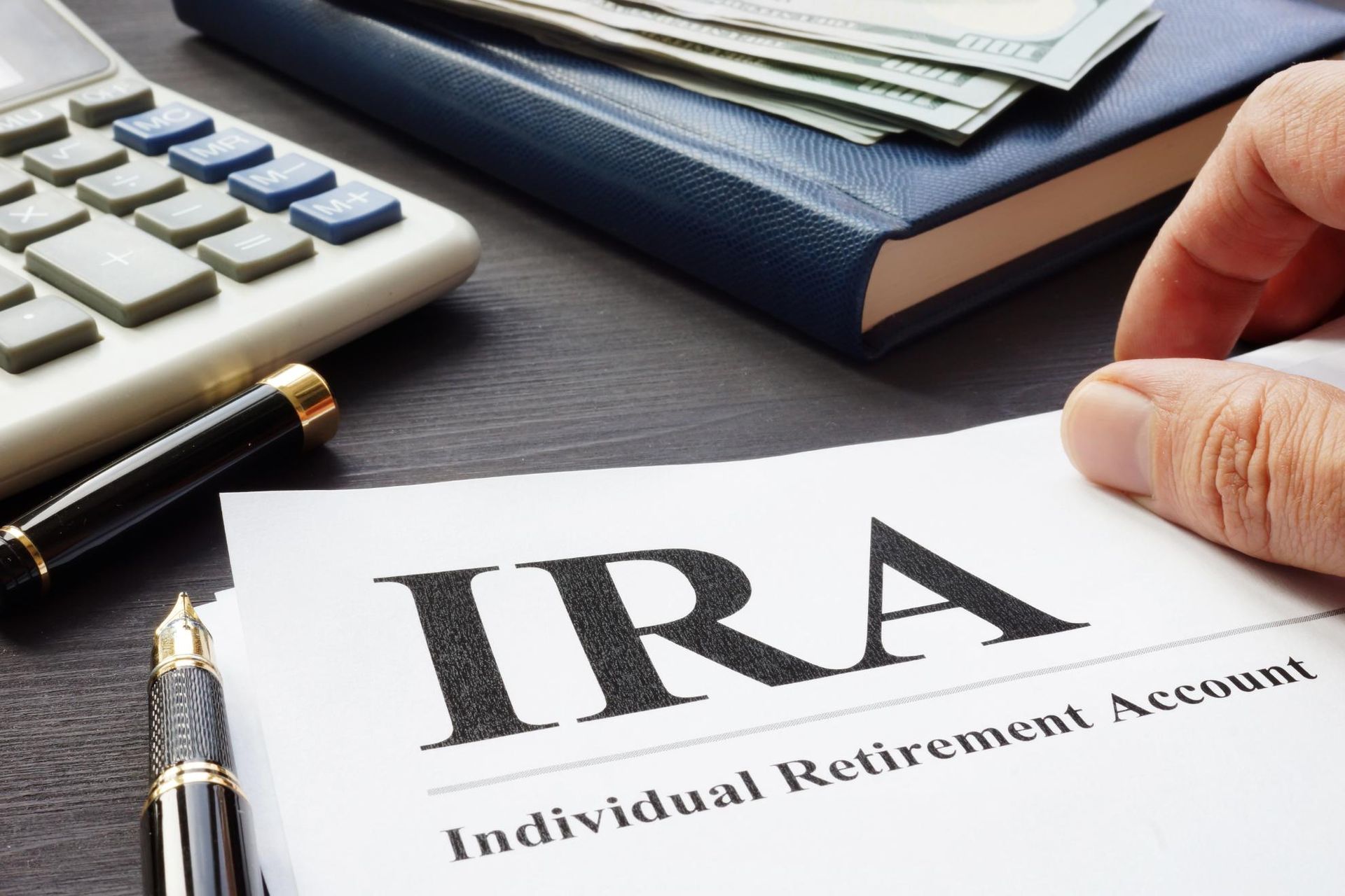 Hand holding IRA document with money, calculator, pen, and blue notebook on a desk.