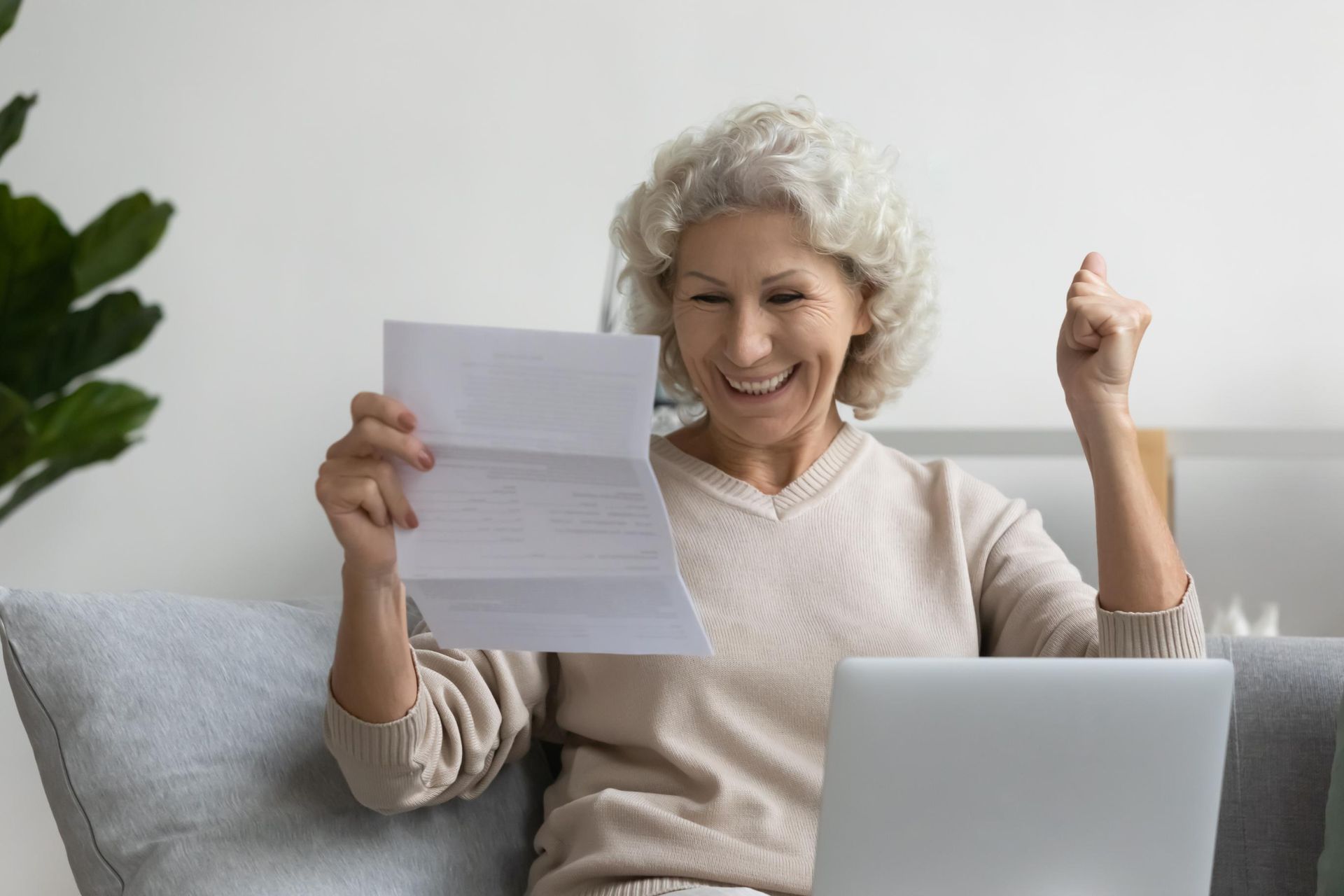 Excited older woman holding a letter, sitting in front of a laptop, clenching fist in joy.