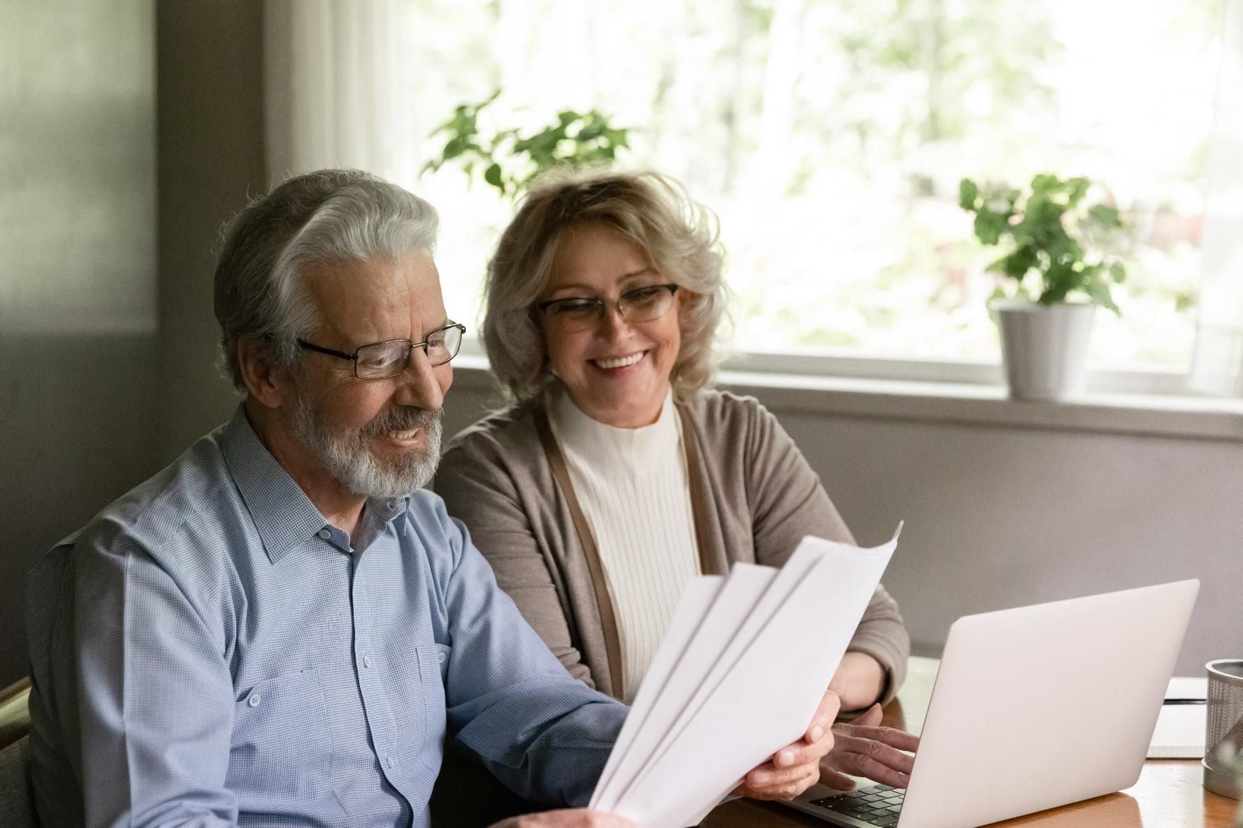 Elderly couple reviewing documents together, smiling near a laptop indoors.
