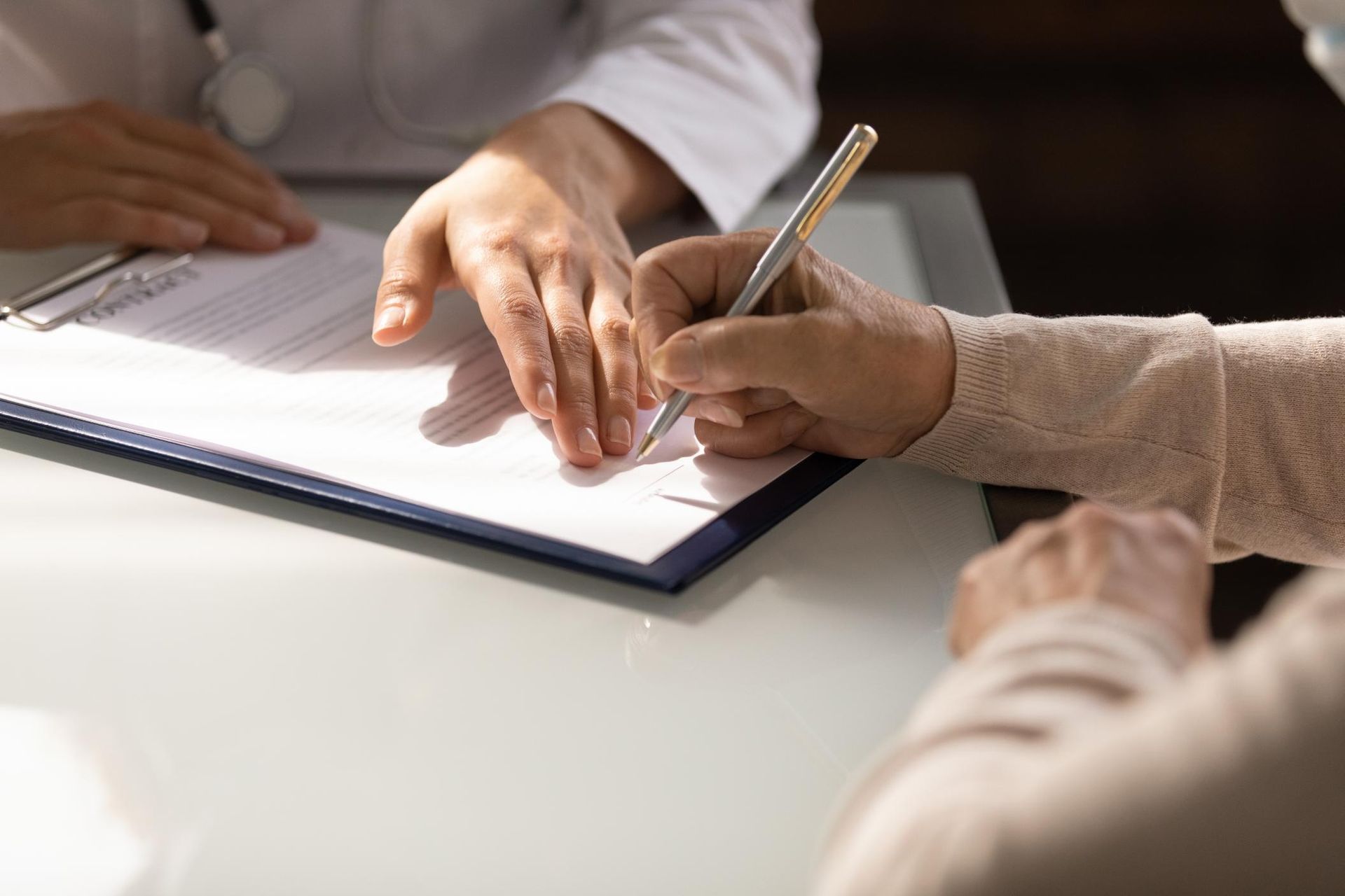 Doctor assisting a patient with signing a document on a clipboard at a desk.