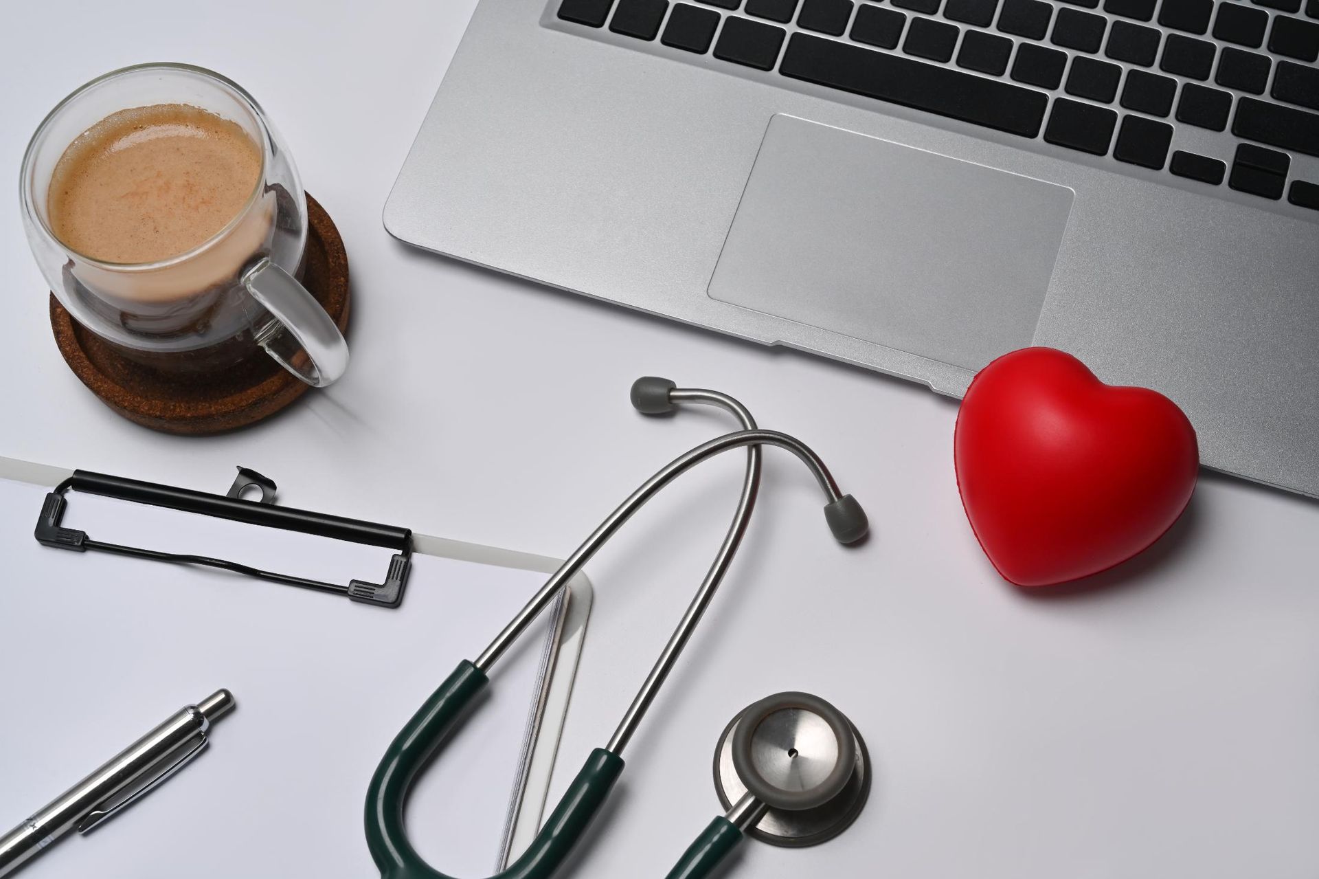 Laptop, stethoscope, red heart, coffee, clipboard, pen on white surface.