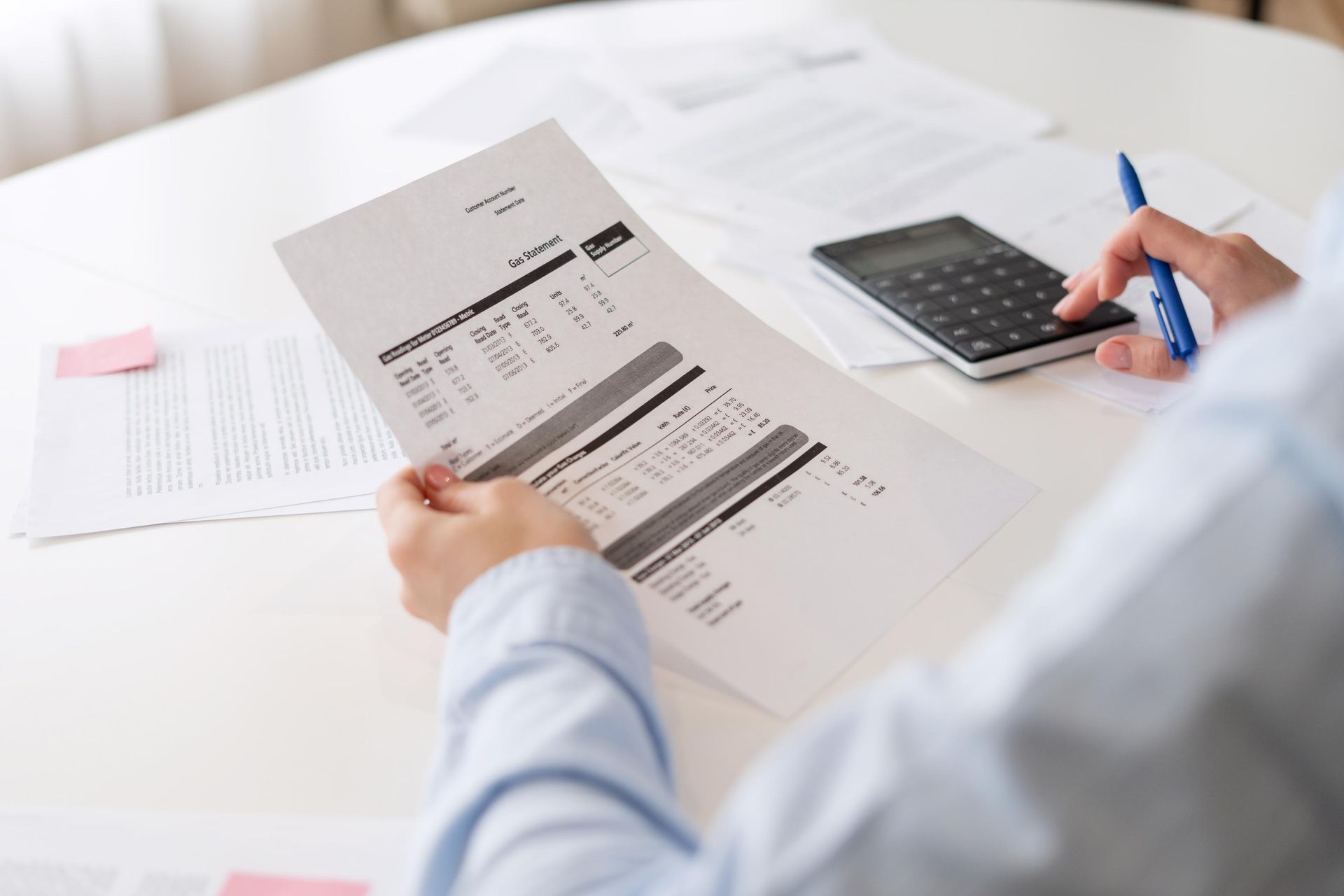 Person in blue shirt uses calculator while reviewing bills at a white table.