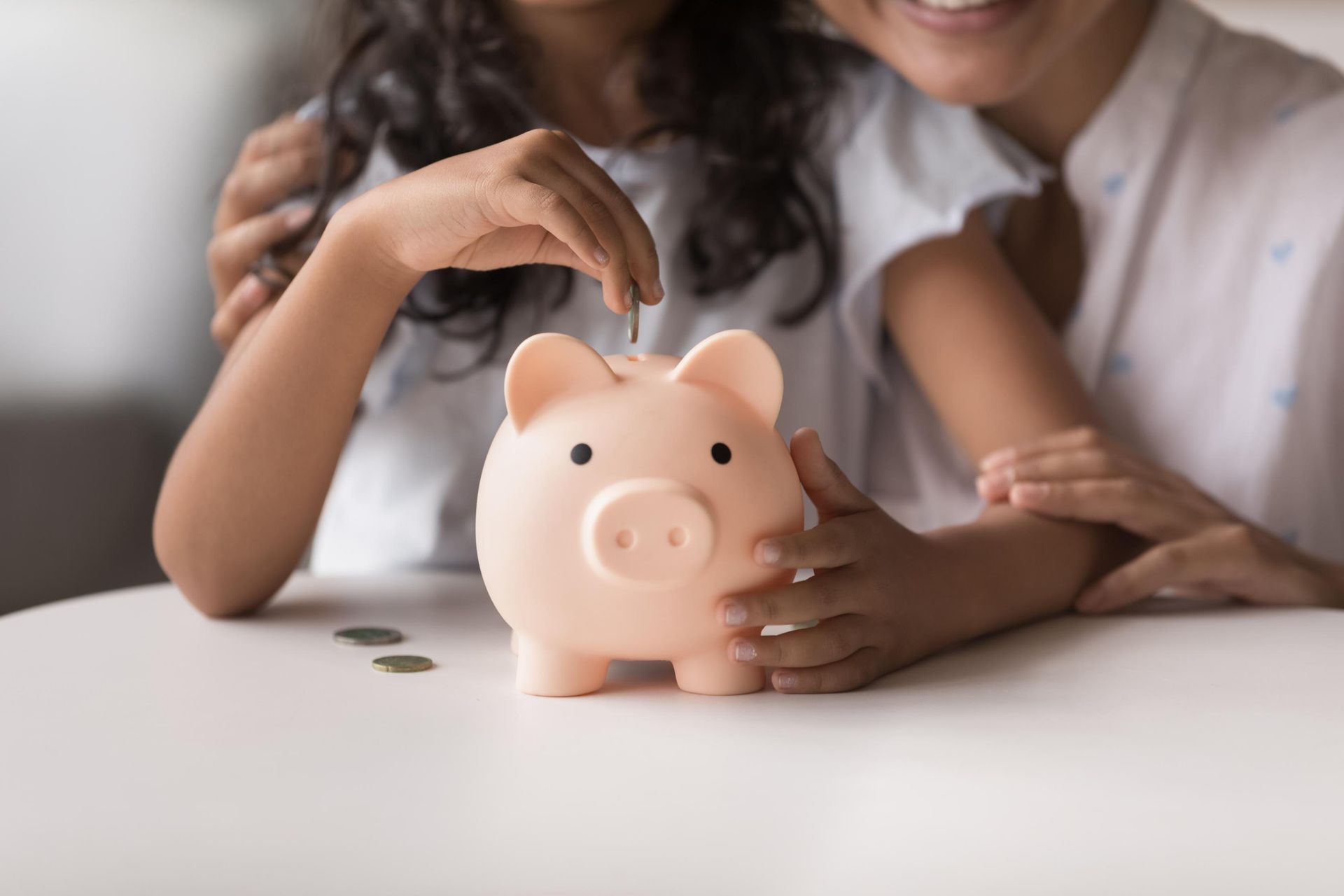 Girl putting coin in piggy bank with adult's arm around her. Light pink bank on white table.