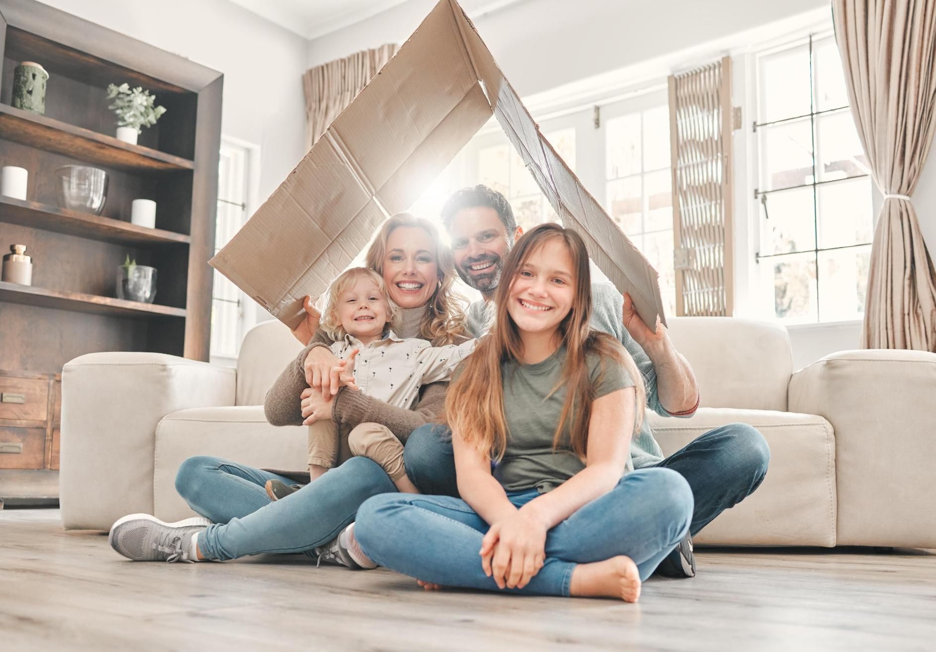 Family of four sits under a cardboard house in a sunlit living room, smiling.