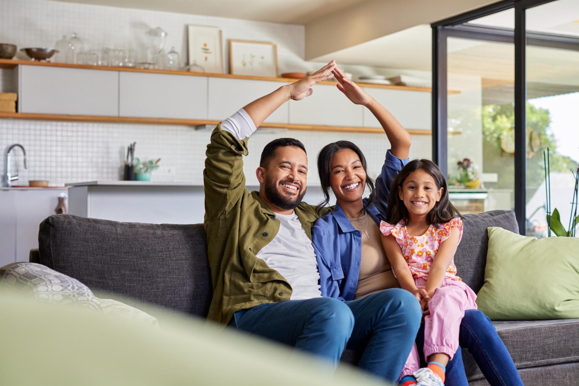 Family of three, smiling on couch, parents forming roof with hands above child in living room.