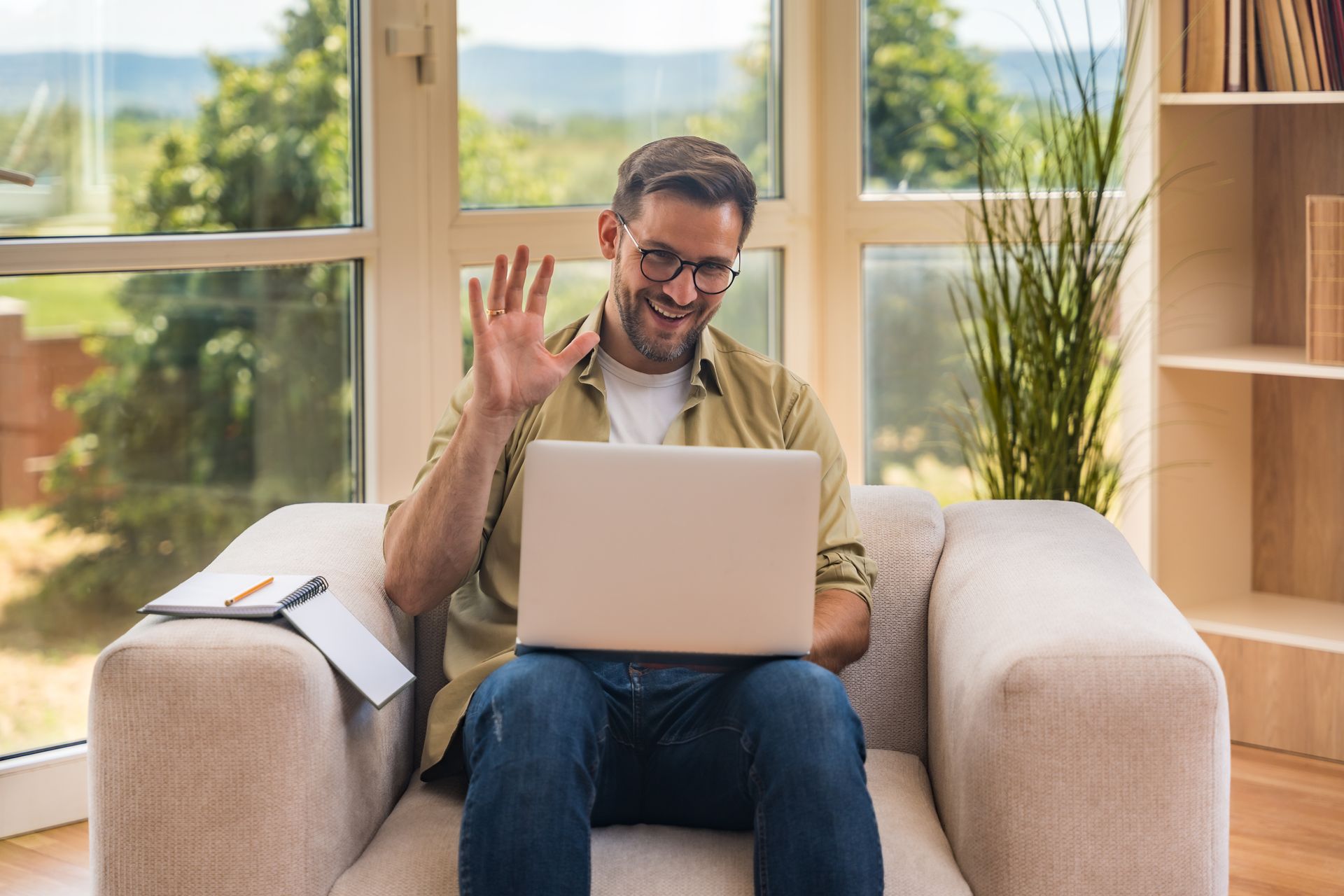 Man waving on a couch while using a laptop in a bright living room with large windows