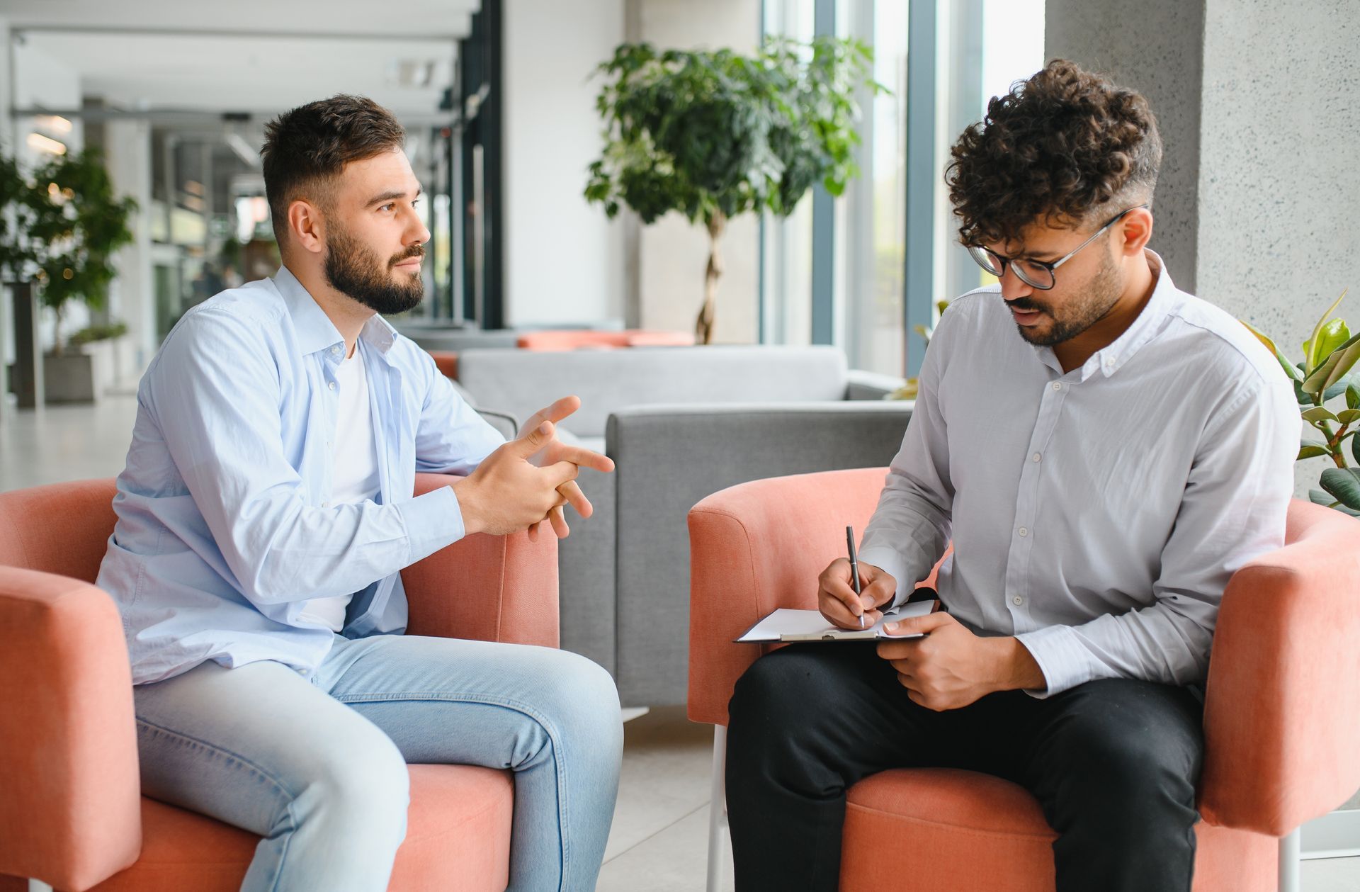 Two people talking in a bright office lounge, one taking notes on a tablet.