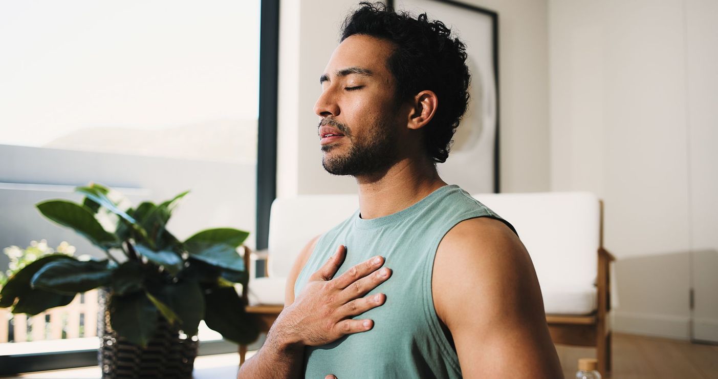 Man sitting indoors with eyes closed, hand on chest, in a calm breathing pose near a window and plant.