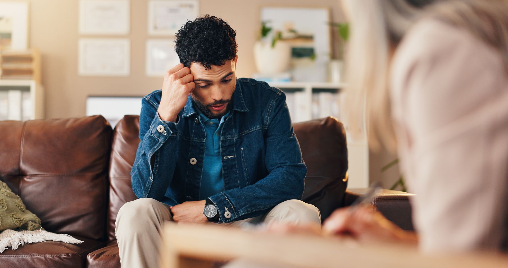 Man seated on a couch, leaning forward with a hand on his forehead, in a cozy living room during a conversation