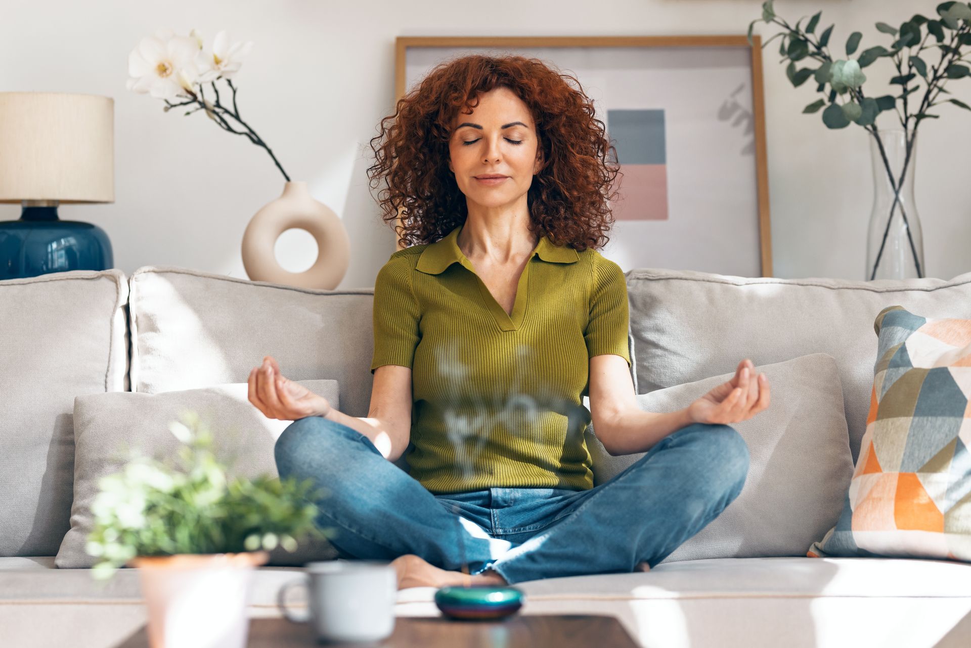 Person meditating cross-legged on a couch in a bright living room, holding hands in a relaxed pose.
