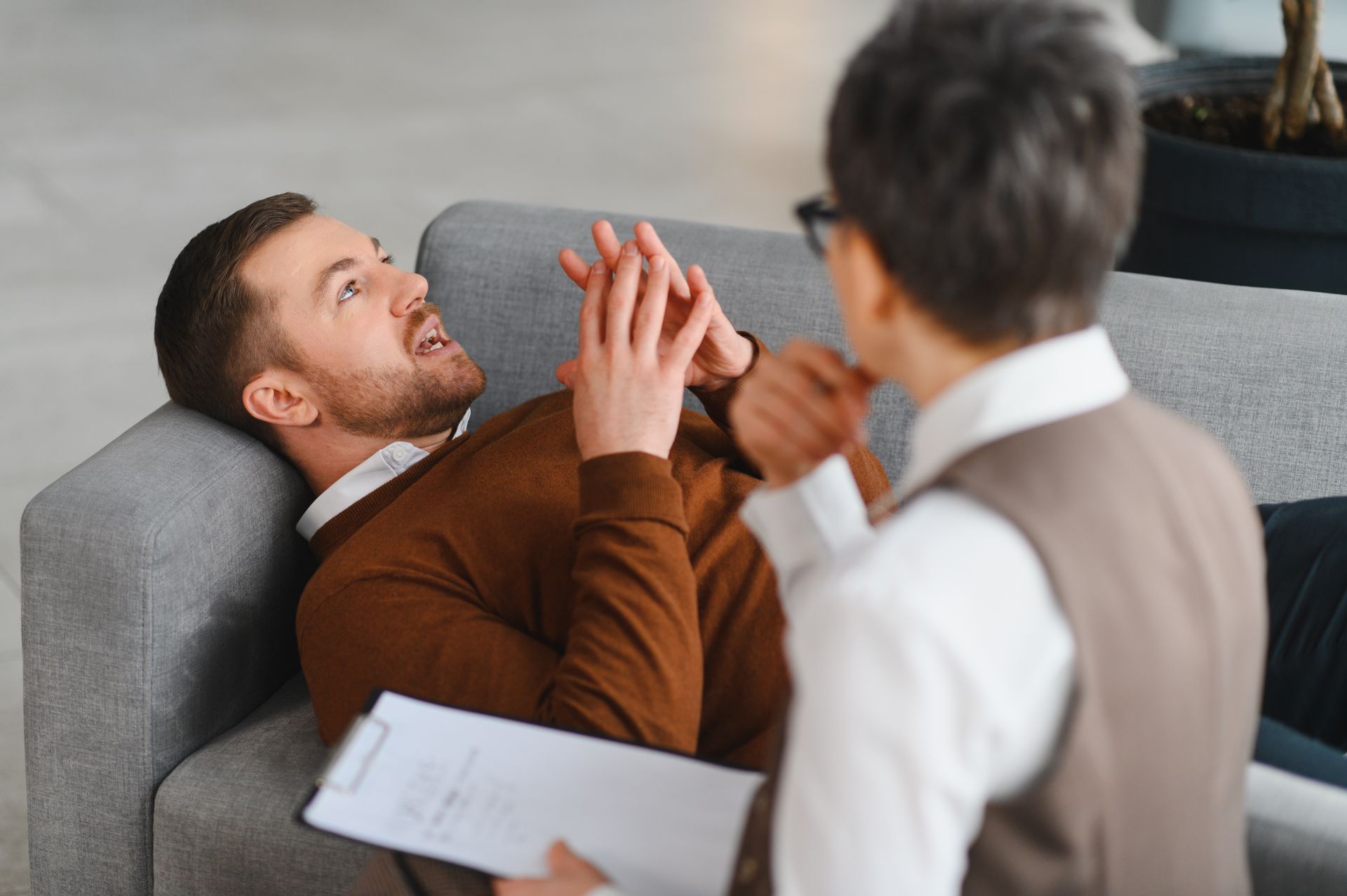 Counselor talking with a reclined client on a gray sofa, holding a clipboard in a bright office