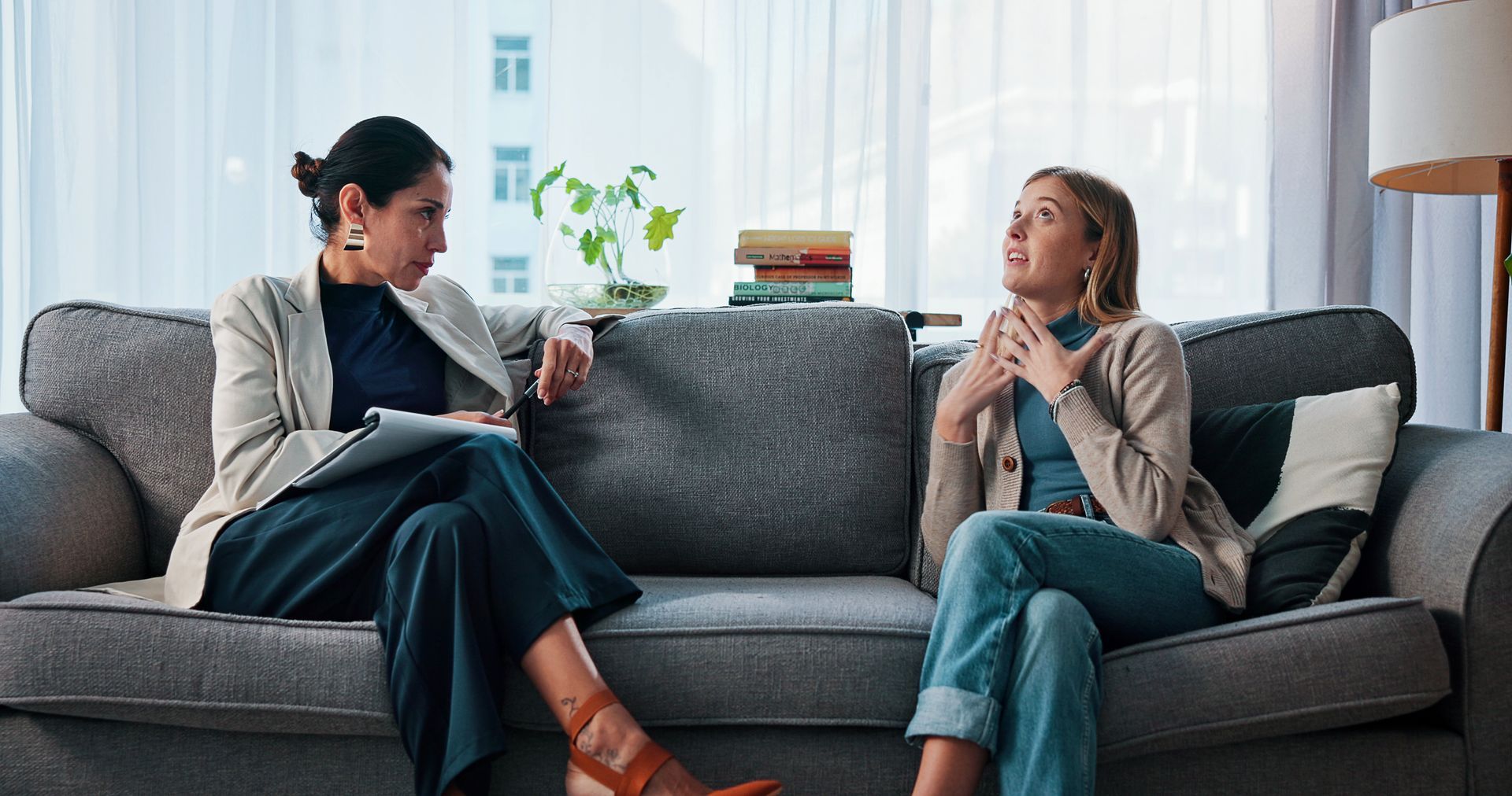 Two people talk on a gray couch in a bright living room, one holding a notebook.