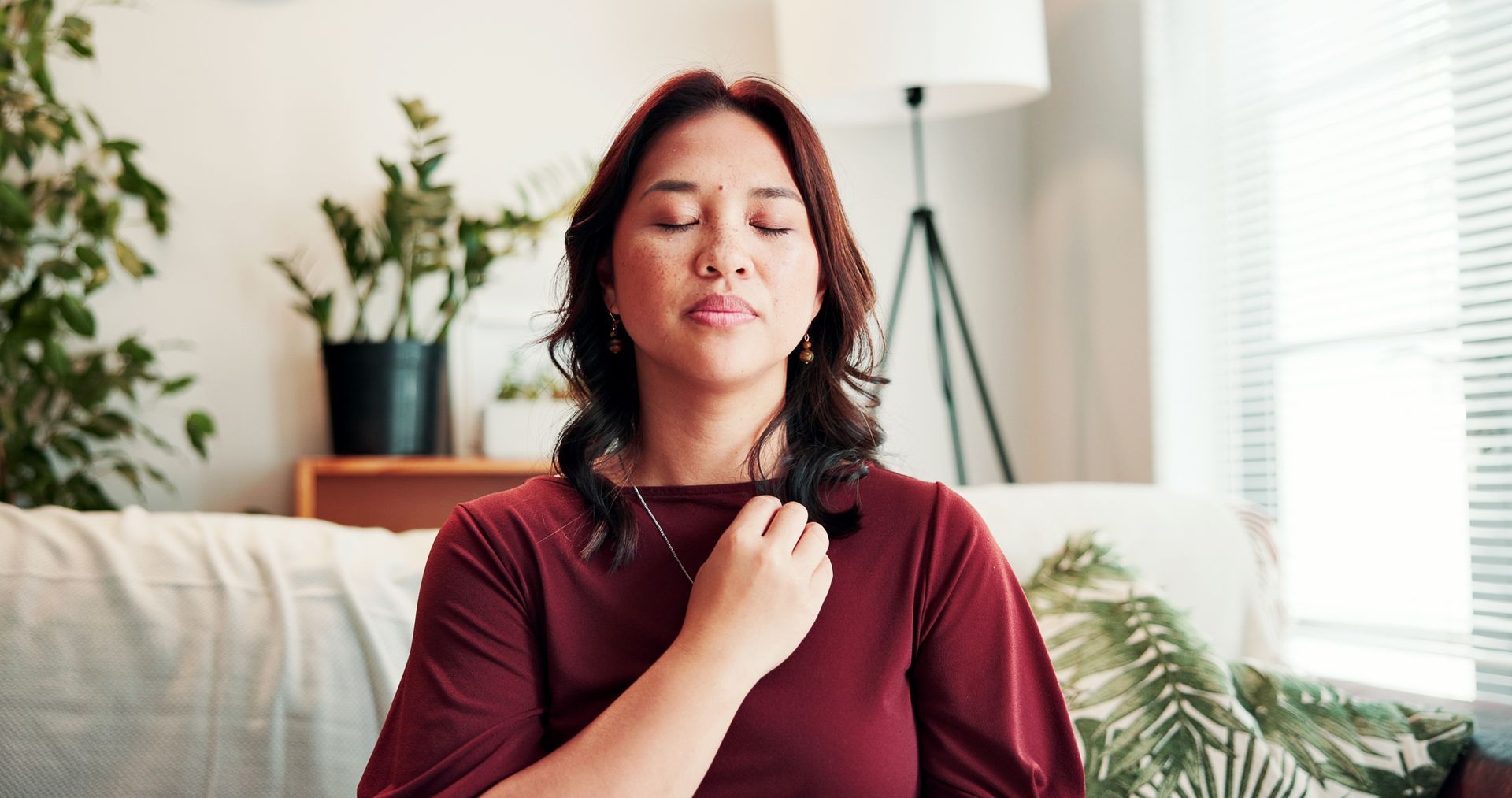 Woman sitting on a couch, eyes closed, touching her throat in a bright living room.