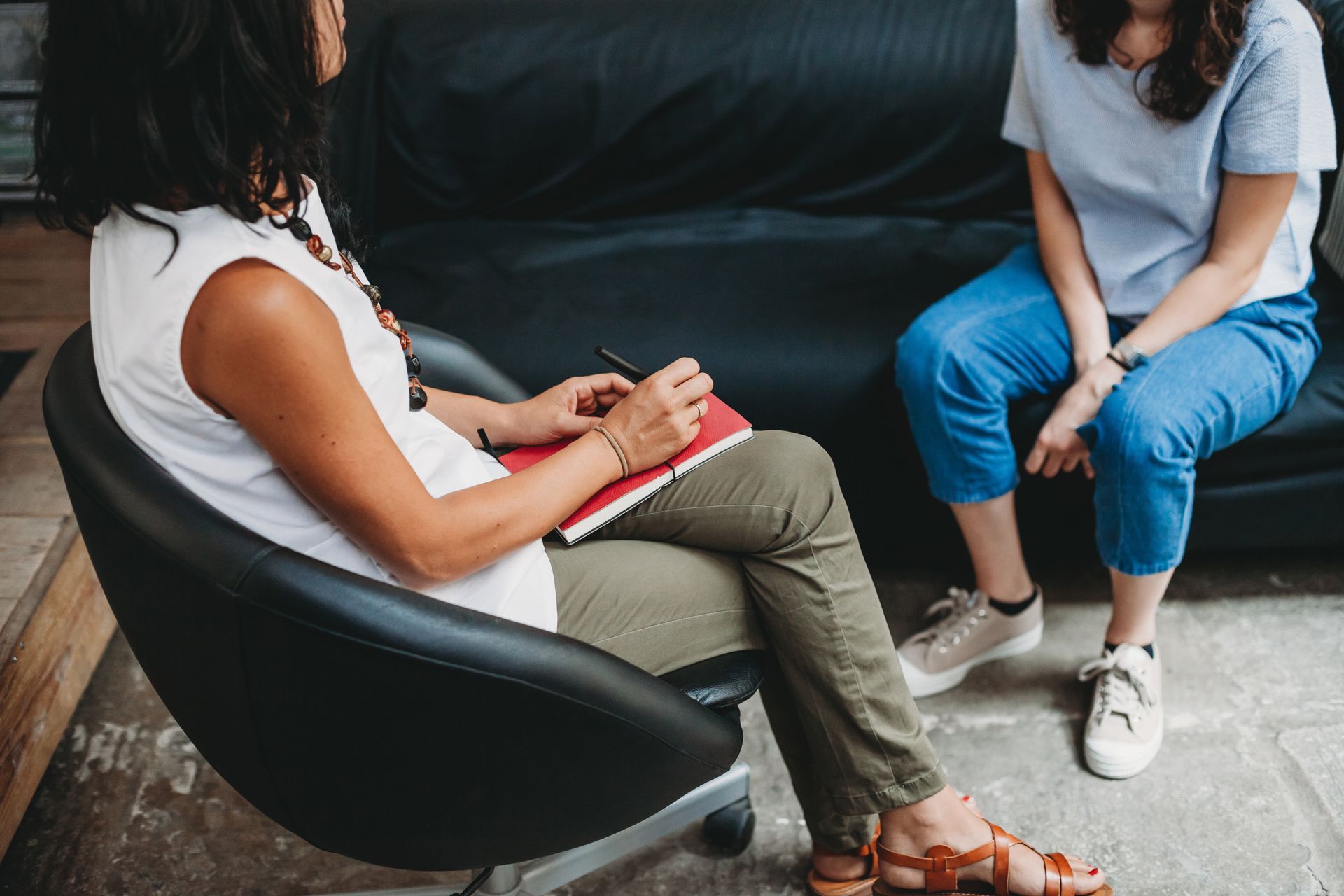 Two people seated indoors in conversation, one holding a notebook on a black chair and the other on a sofa.