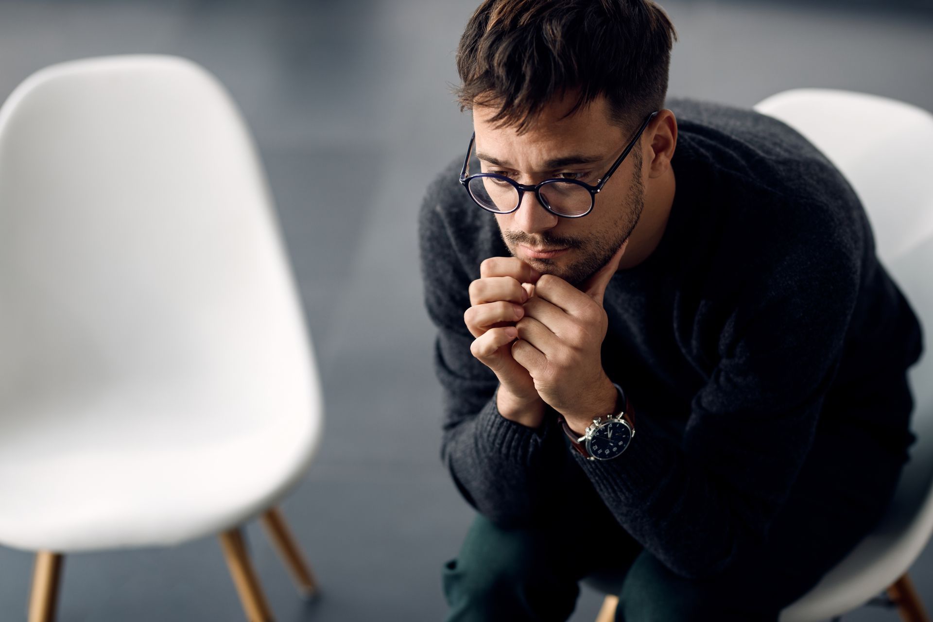 Man sitting pensively between two white chairs, hands clasped near his face in a modern room