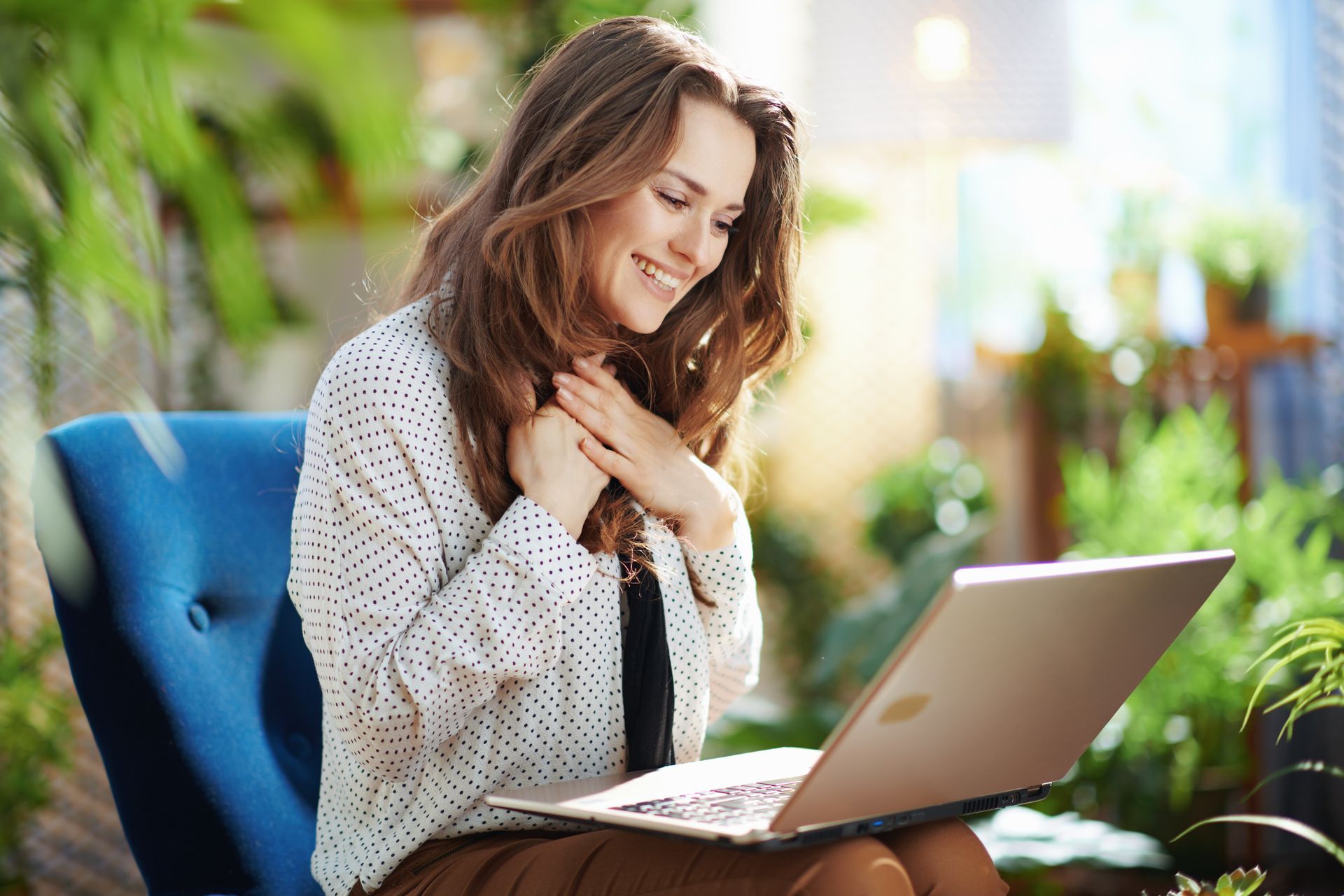 Woman smiling on a blue chair, using a laptop in a bright room with plants.