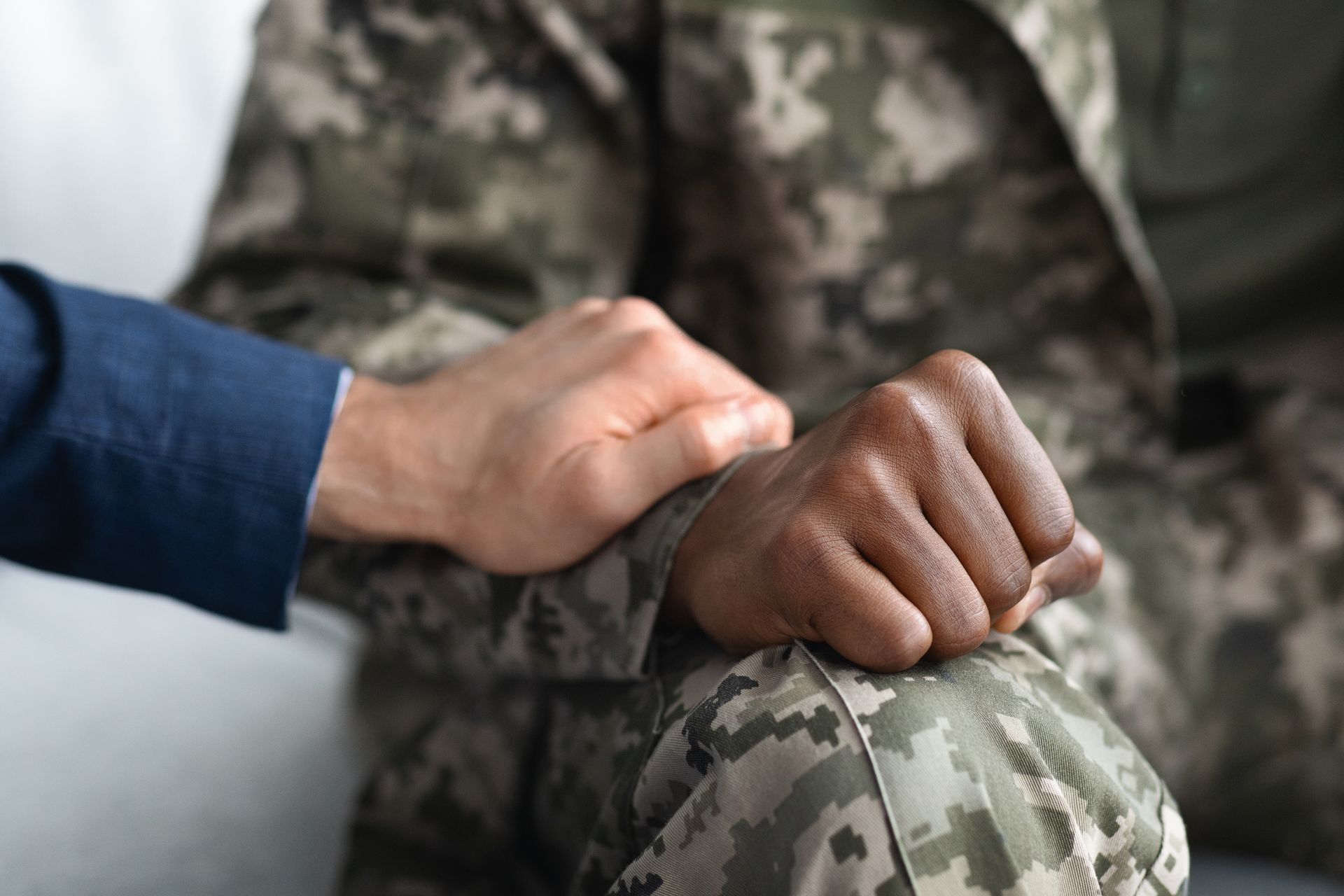 A hand rests on a soldier’s camouflaged knee in a supportive gesture.