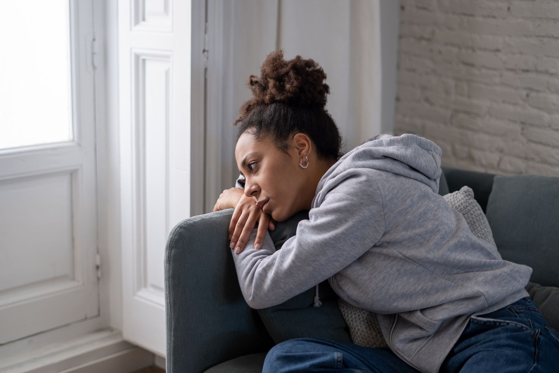 Person in a gray hoodie sitting curled on a couch by a window, resting their head on their hands.
