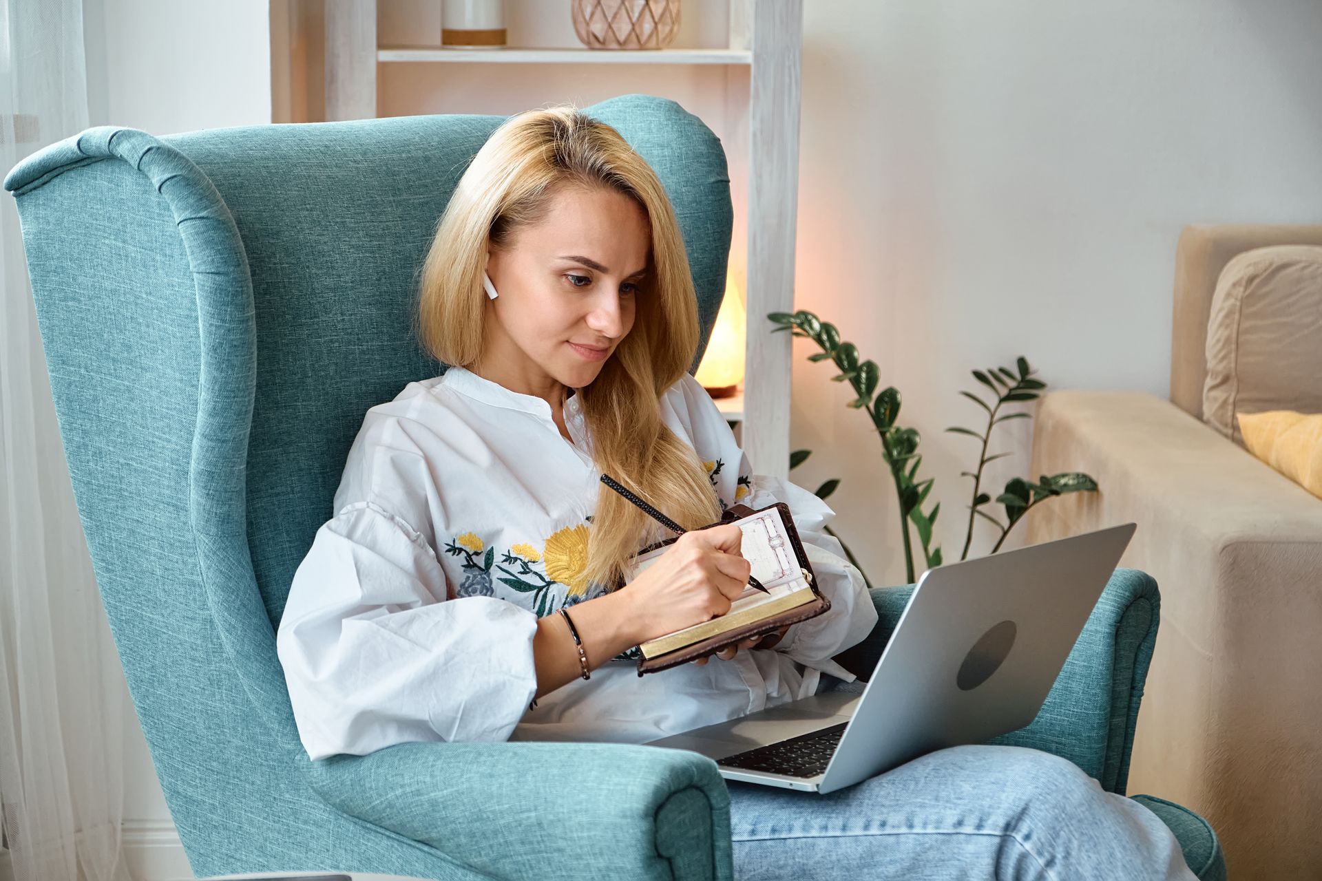 Woman in a teal armchair using a laptop and writing in a notebook at home