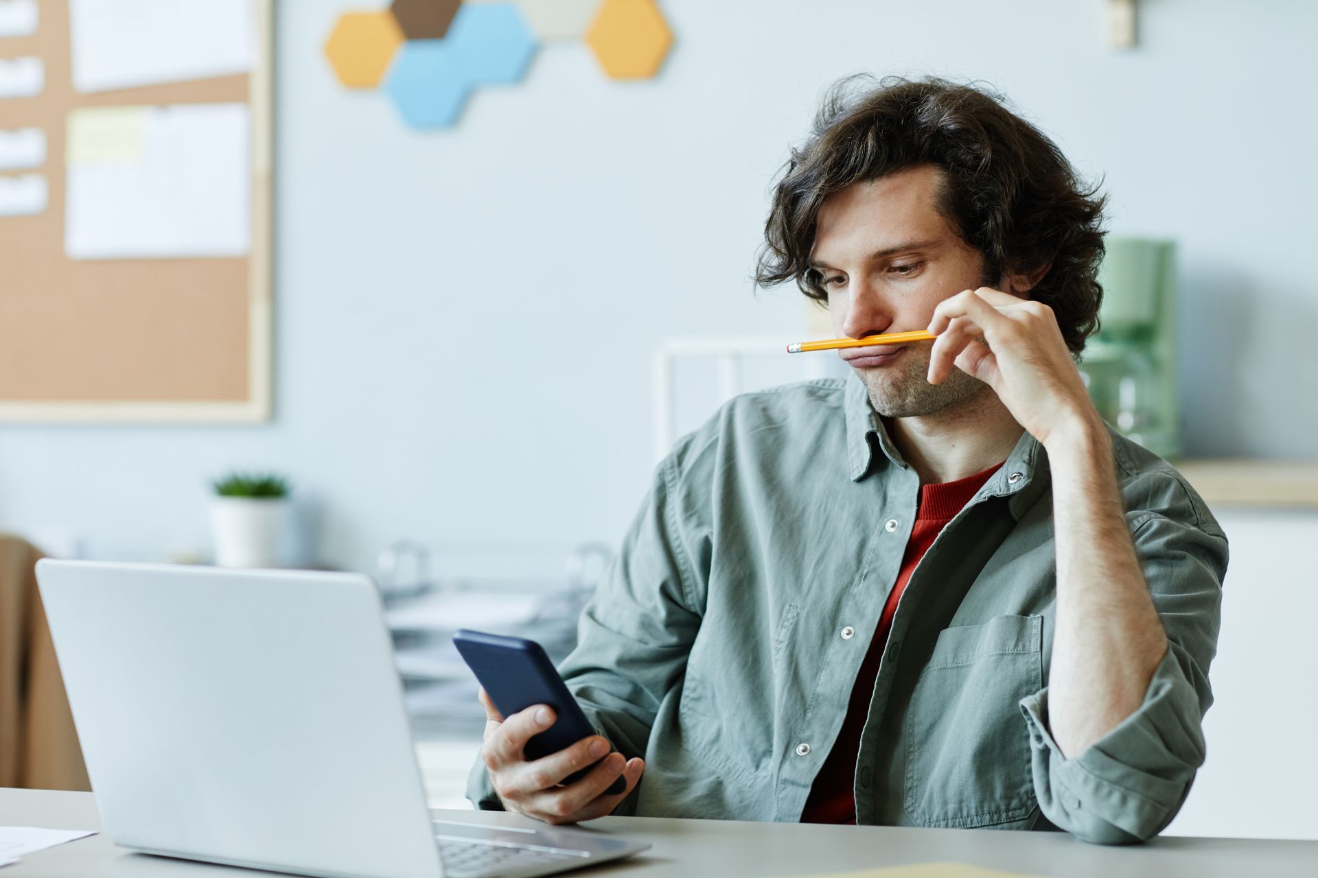 Person holding a phone and pencil at a desk with a laptop in a bright office