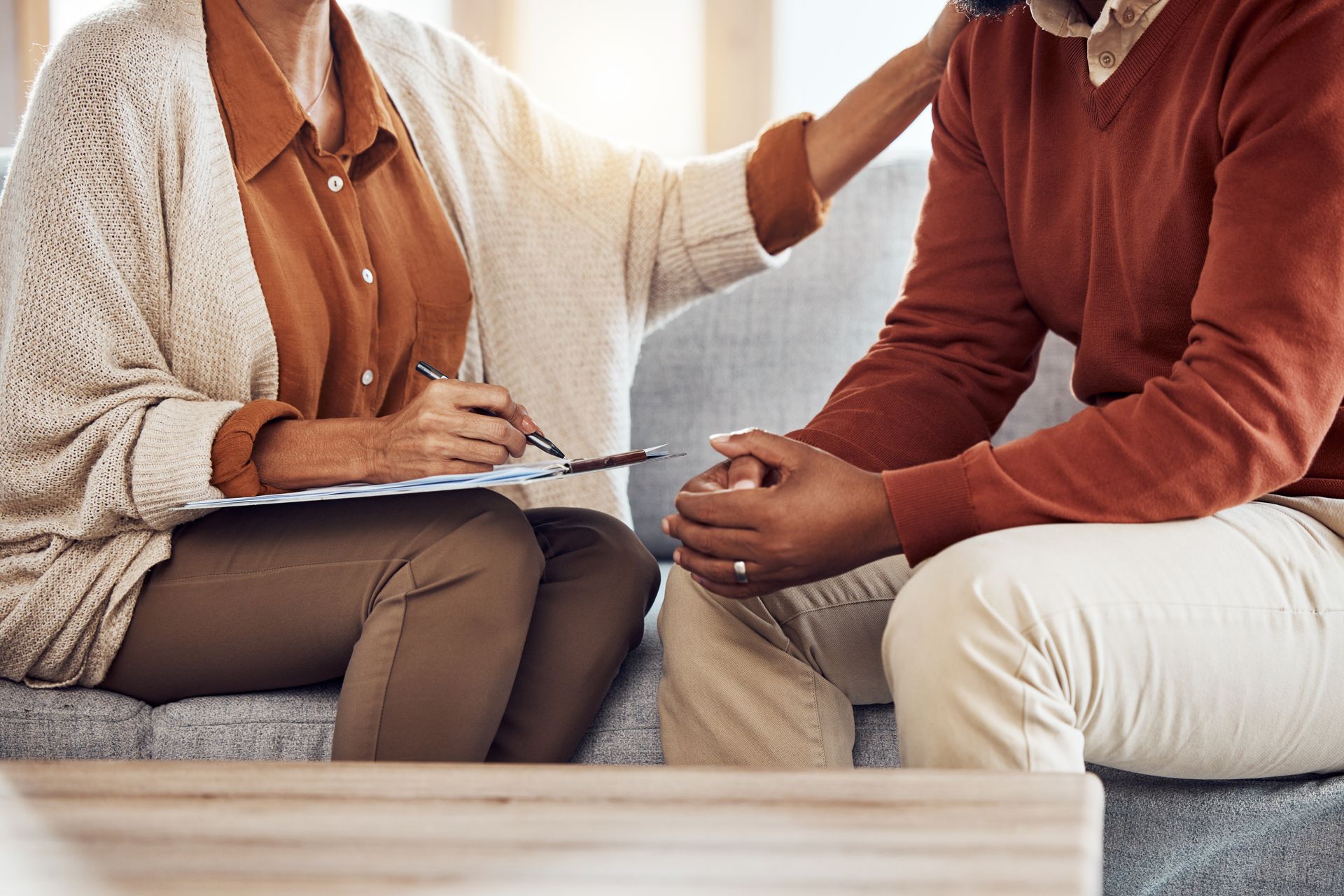 Two people sit on a couch in a counseling session, one taking notes on a clipboard.