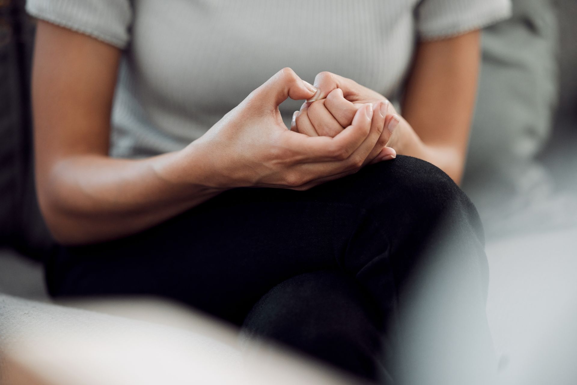 Hands clasped in a calming gesture on a seated person’s lap, wearing a gray shirt and black pants.