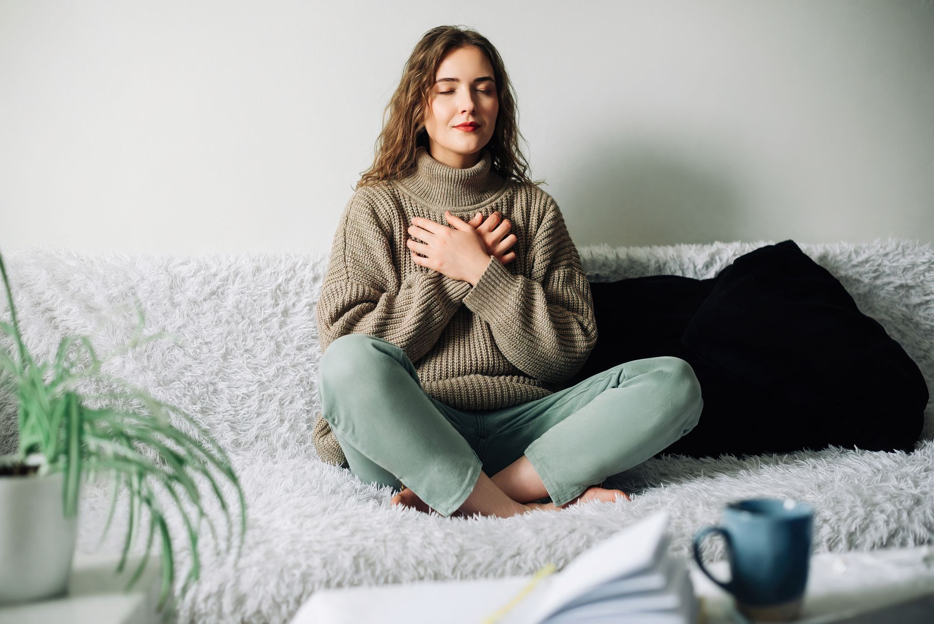 Woman sitting cross-legged on a white couch, hands over heart, in a cozy living room
