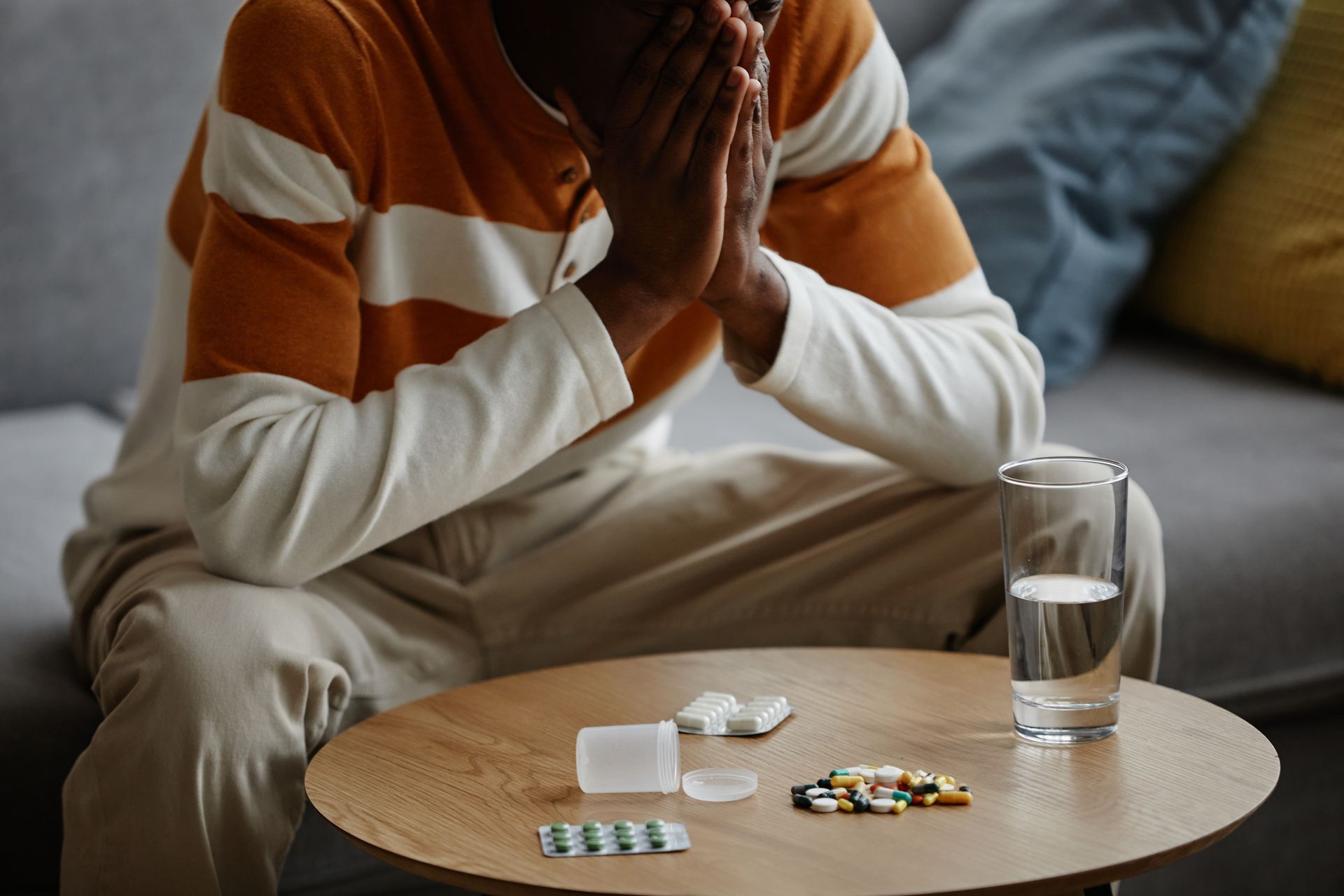 Person sitting at a table with pills, holding their face in distress beside a glass of water