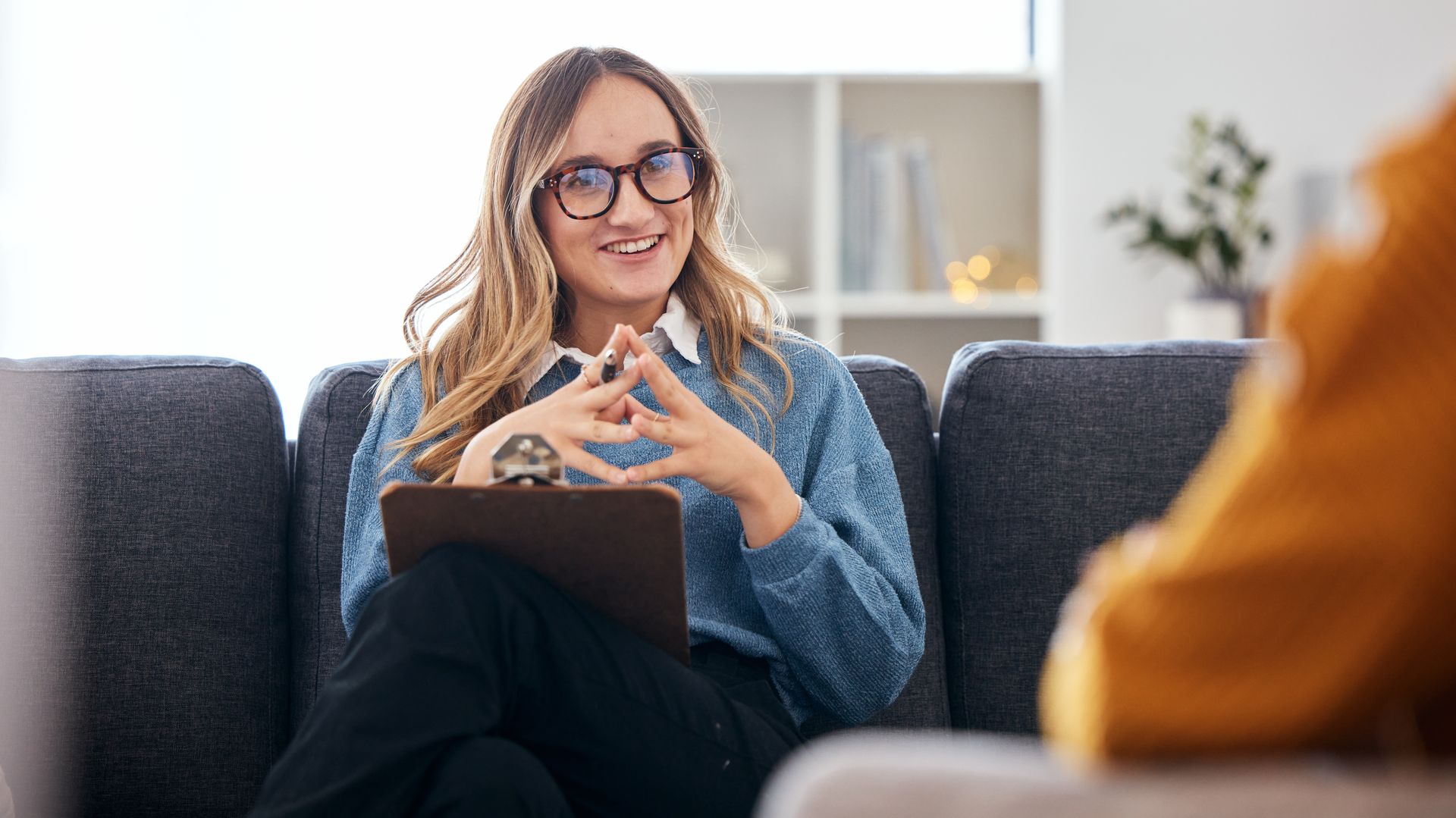 Woman with glasses speaking to a counselor on a sofa, holding a clipboard in a bright office
