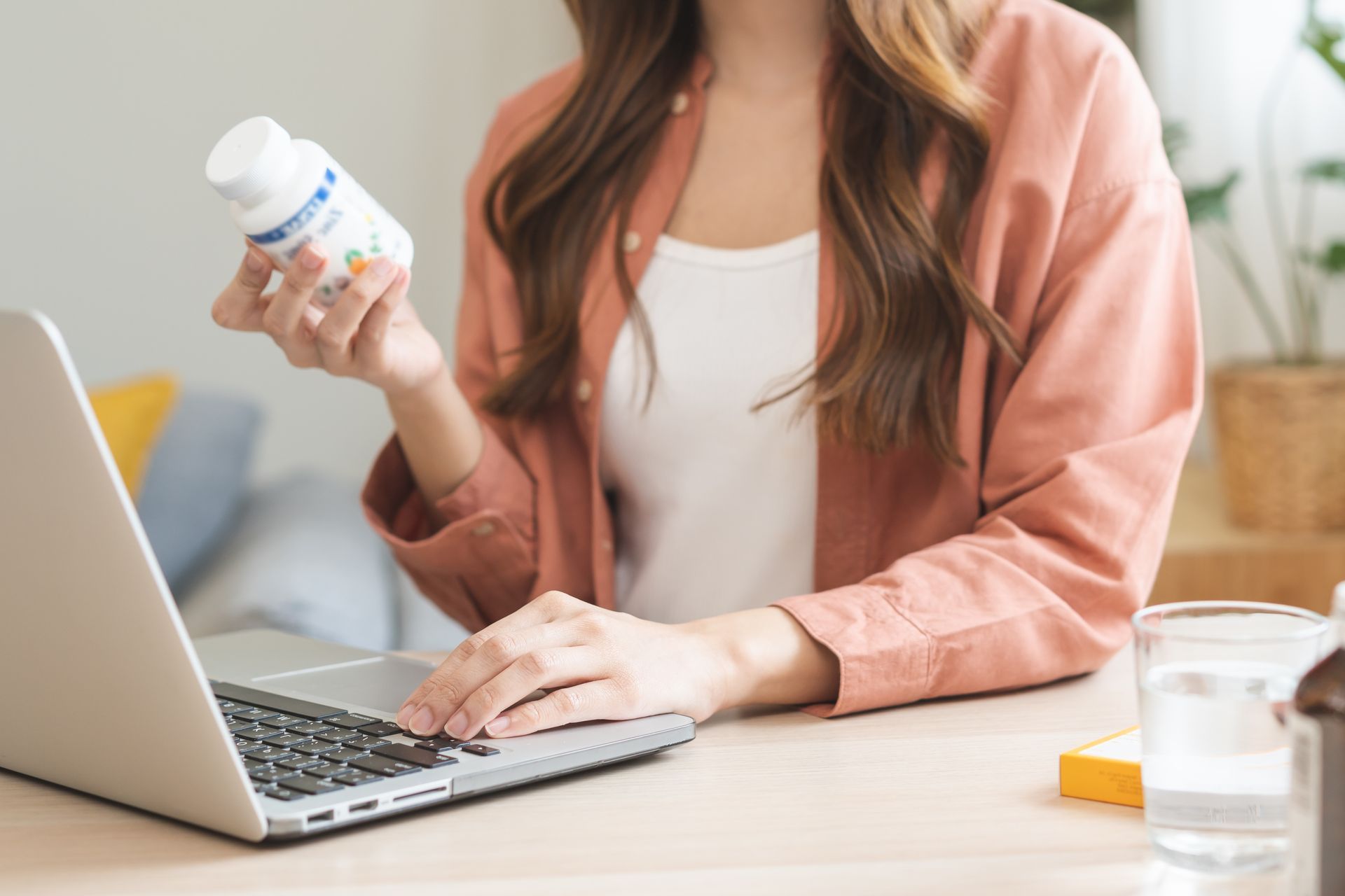 Person holding a pill bottle while using a laptop at a desk in a bright room