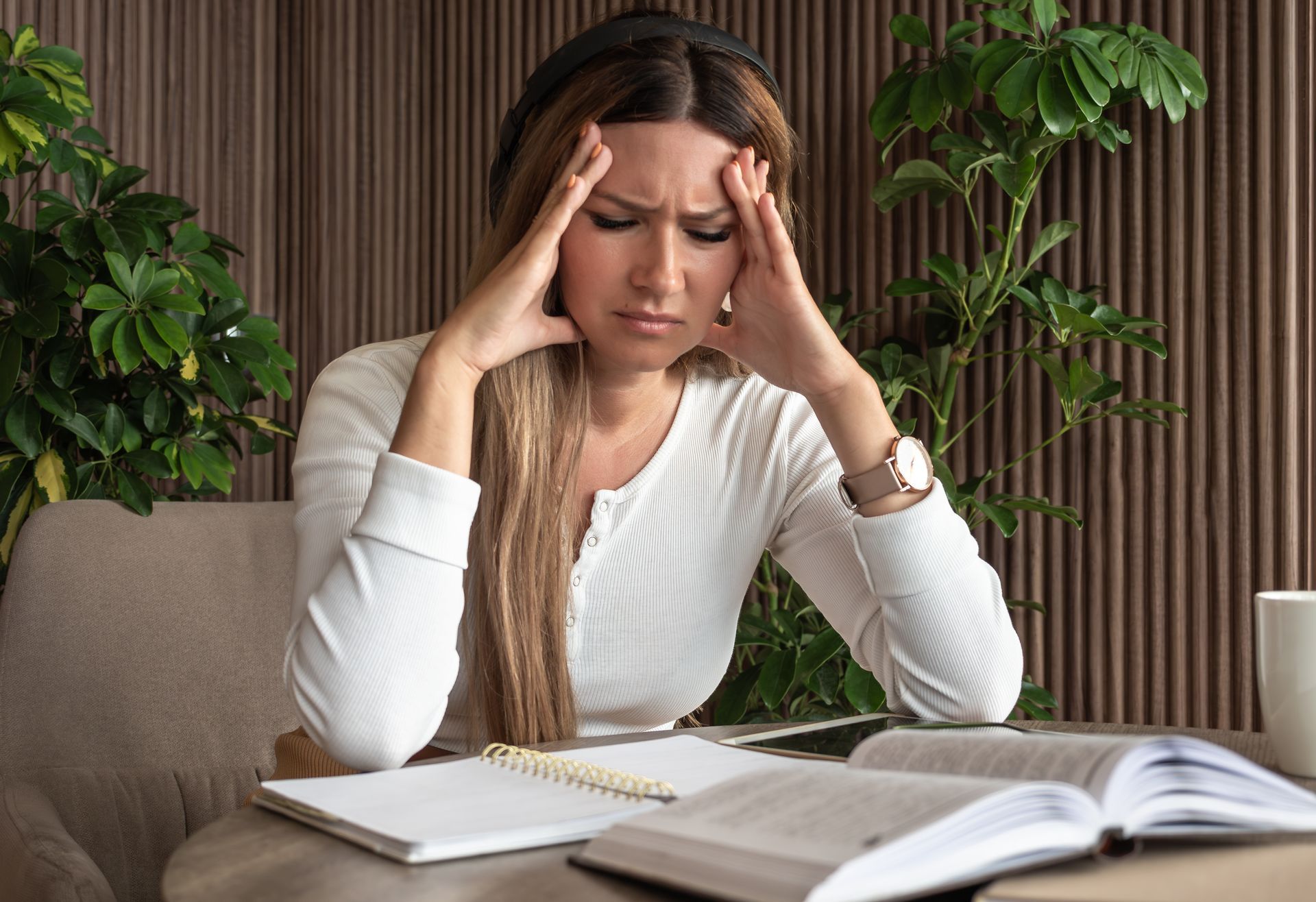 Woman at a desk, holding her temples with a stressed expression beside open books and papers.