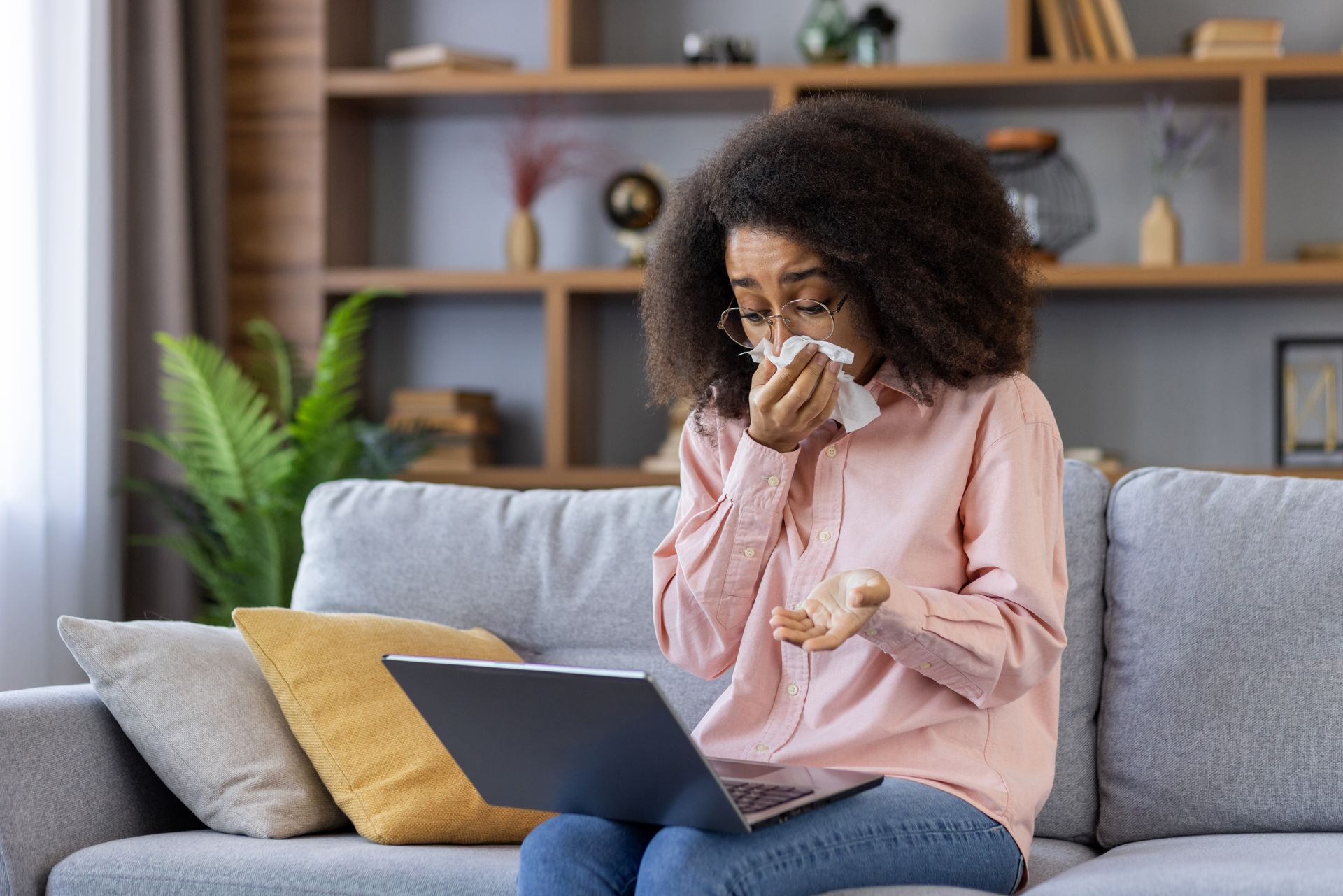 Person on a couch uses a tissue while looking at a laptop in a living room