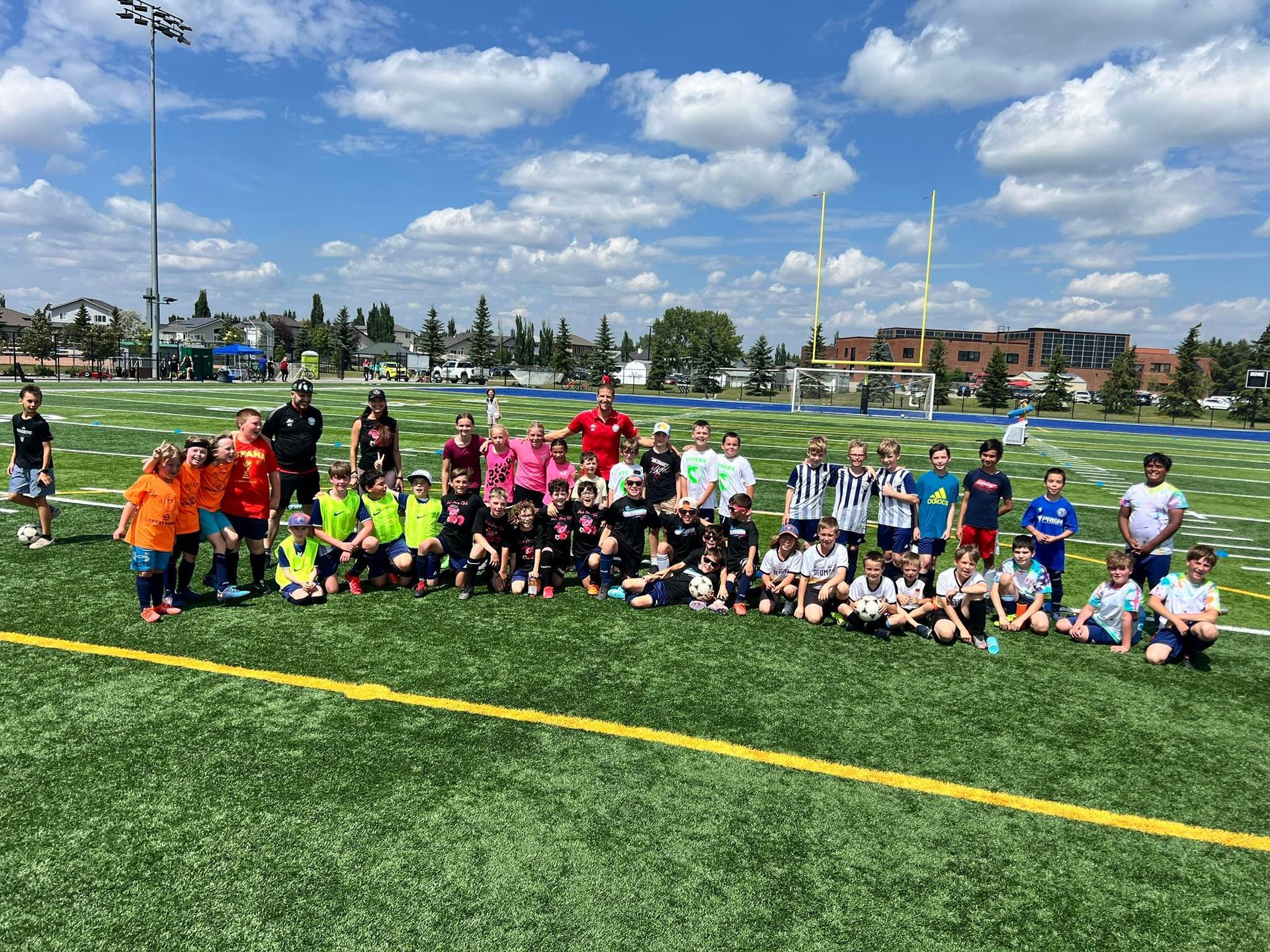 Youth soccer teams pose on a sunny field, wearing bright jerseys in orange, pink, green, and blue.