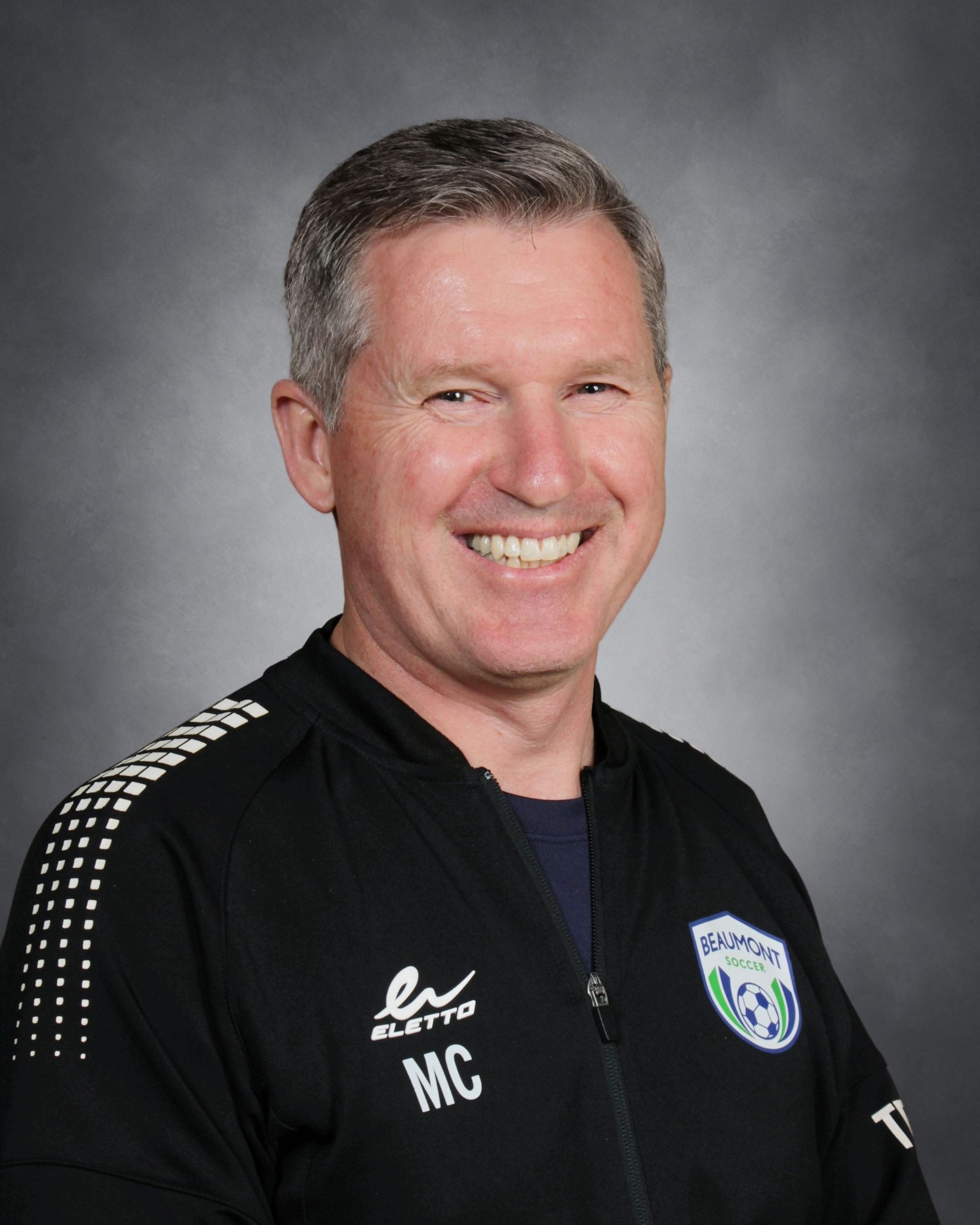 Smiling person in a blue sports jacket with logos, posed against a gray studio background