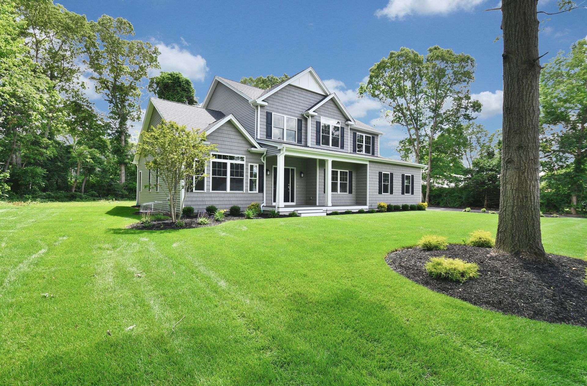 Gray house with black shutters on a large green lawn, surrounded by trees on a sunny day.
