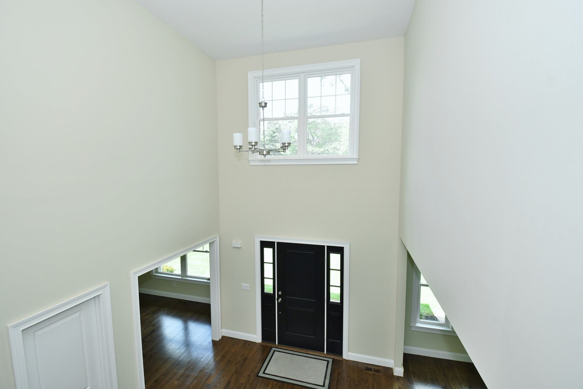 High-angle view of a home's entryway with a dark door, chandelier, and high window. Light-colored walls and hardwood floors.