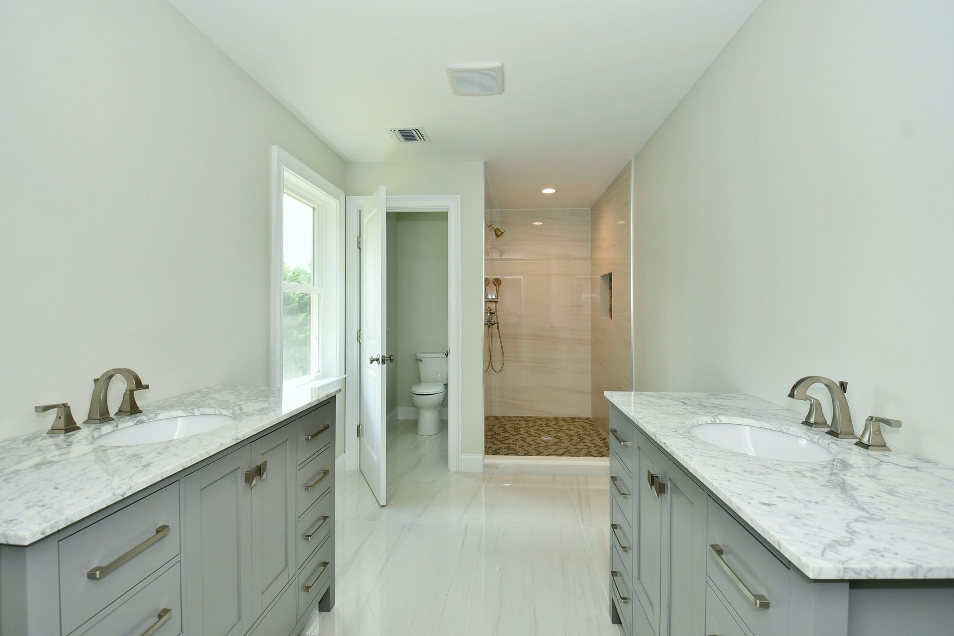 Bathroom with gray double vanity, white countertops, and walk-in shower. A window and open door lead to the toilet.
