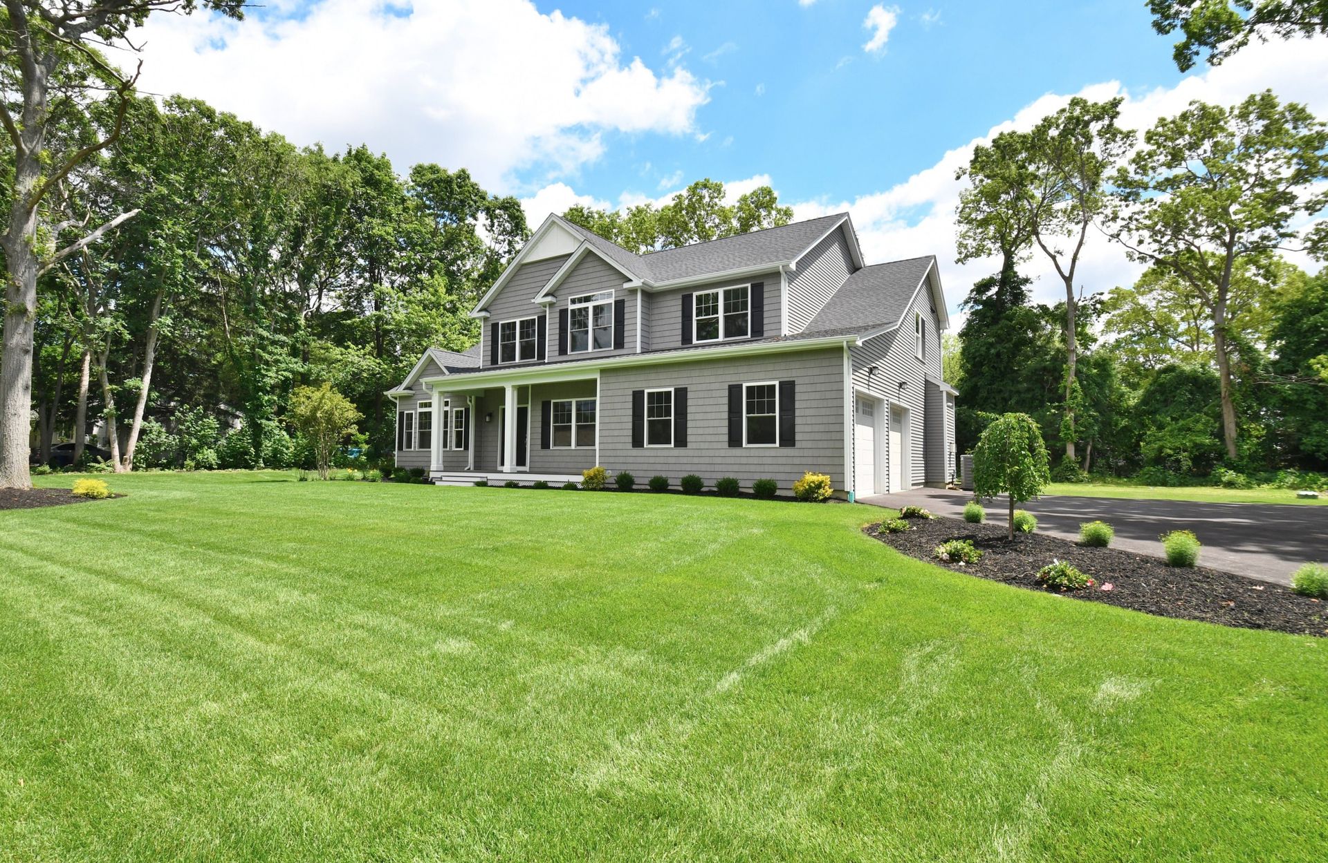 Two-story gray house with black shutters, a white porch, and a large green lawn, trees in the background.