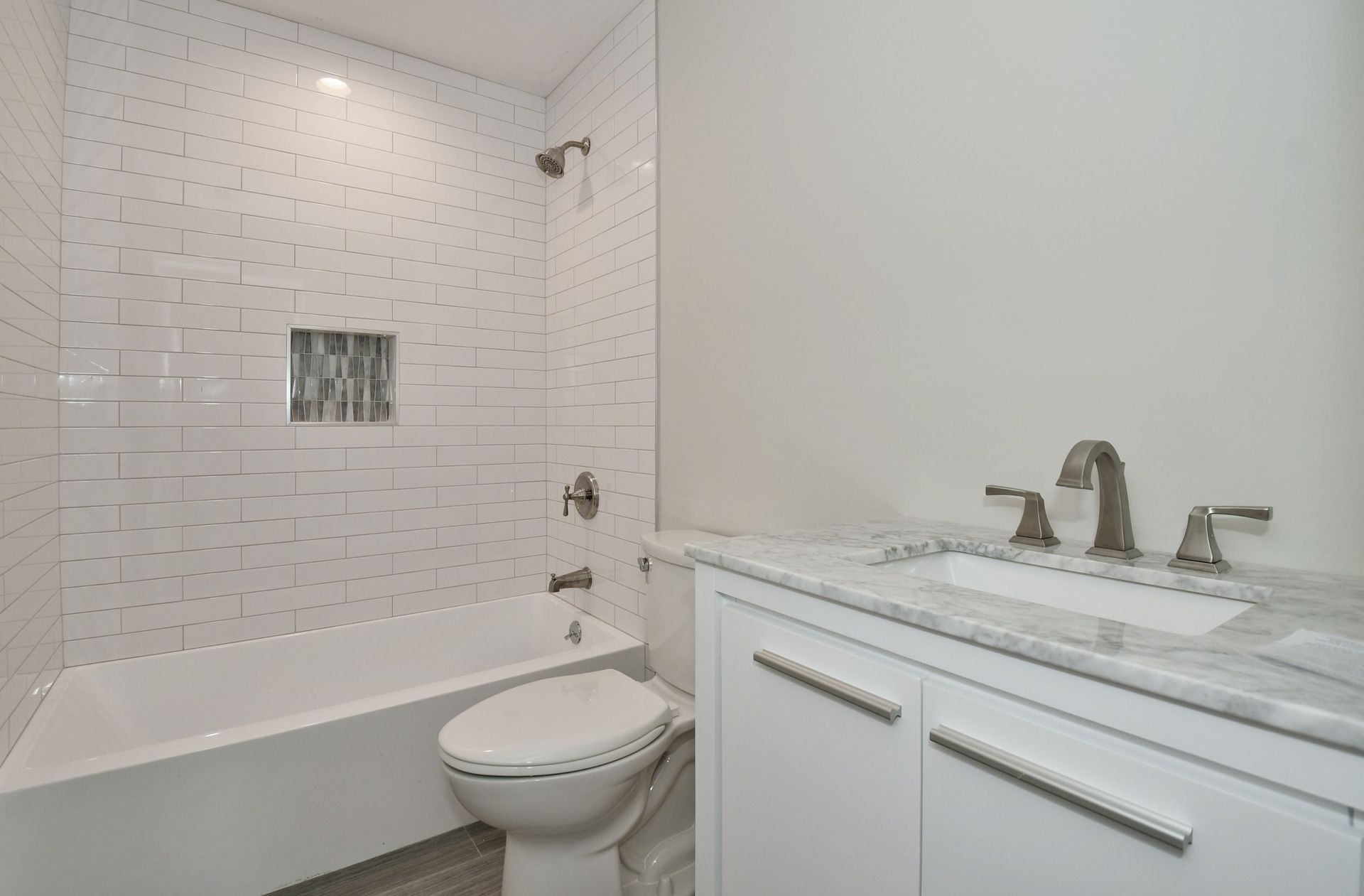 White bathroom with a bathtub, toilet, vanity, and textured tile wall. A silver faucet and showerhead are visible.
